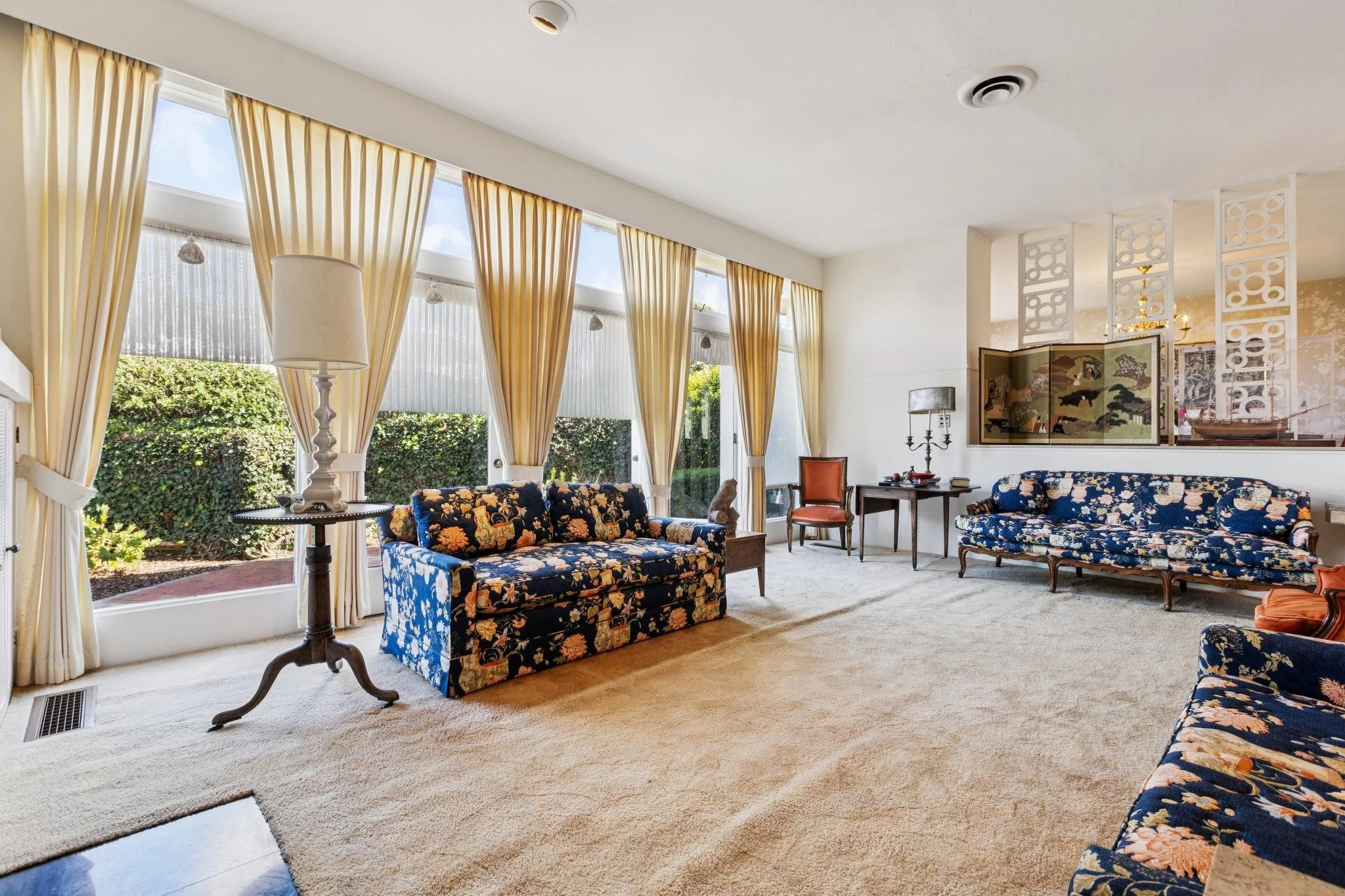 Living room with large windows, yellow curtains, floral patterned sofas, a floor lamp, a wooden side table, and decorative room divider.