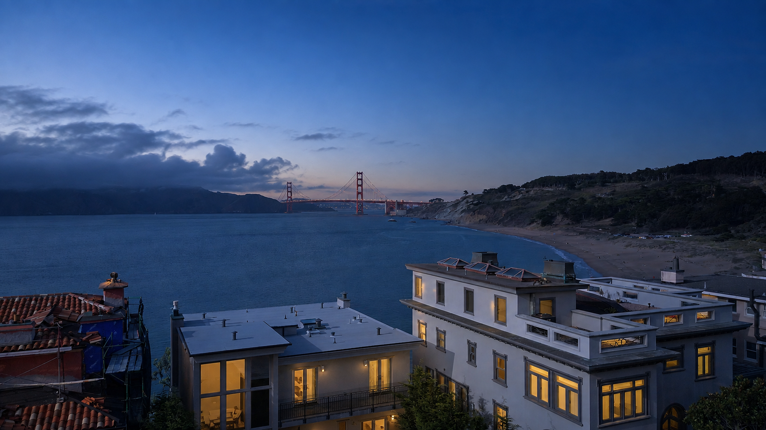Nighttime view of the Golden Gate Bridge over the water, with a partly cloudy sky and nearby residential buildings with lit windows.