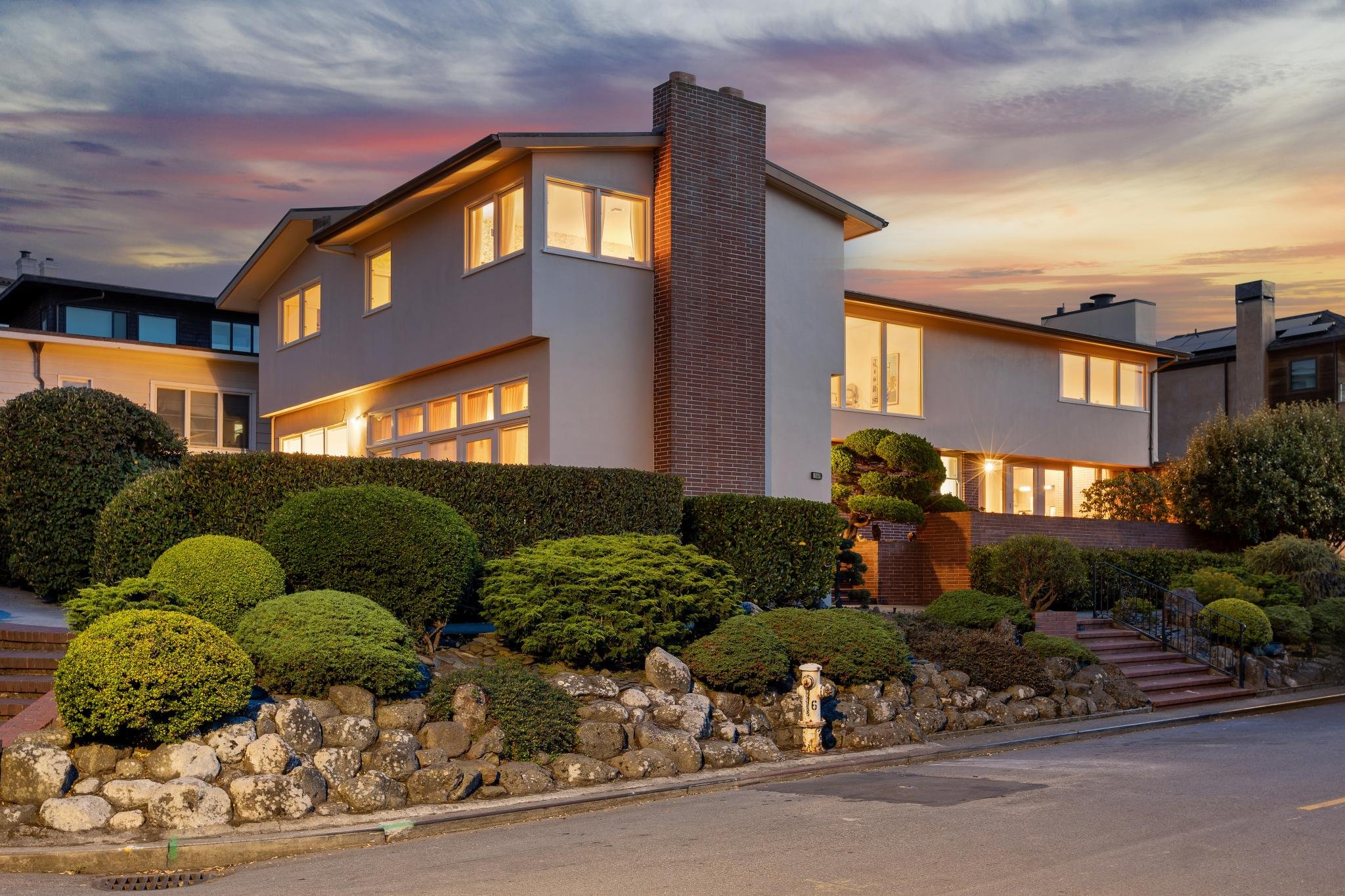 A modern two-story house with large windows, an exterior brick chimney, and lush landscaped bushes, during sunset with a colorful sky.