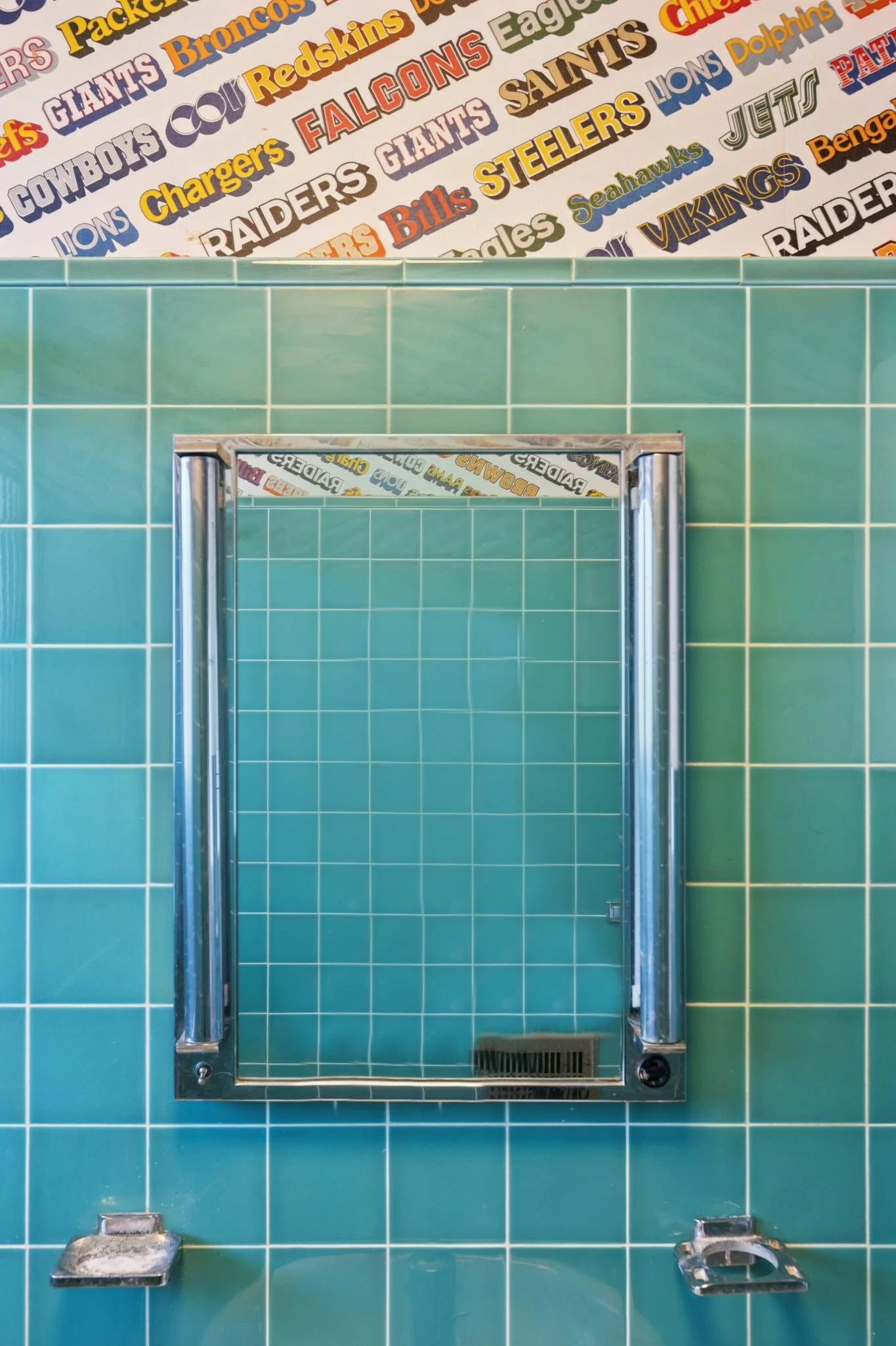 A close-up of a public shower with teal tiles and a metal frame, with sports team posters featuring team names and logos in the background.