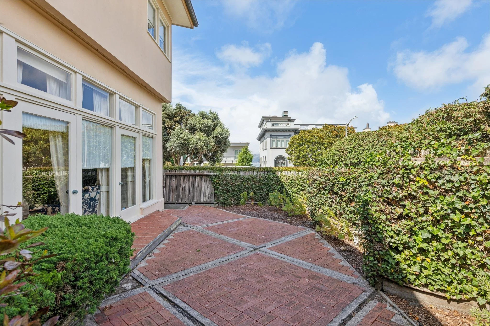 A backyard patio with red brick paving stones, surrounded by green bushes and trees, with a view of neighboring white houses and a blue sky with scattered clouds.