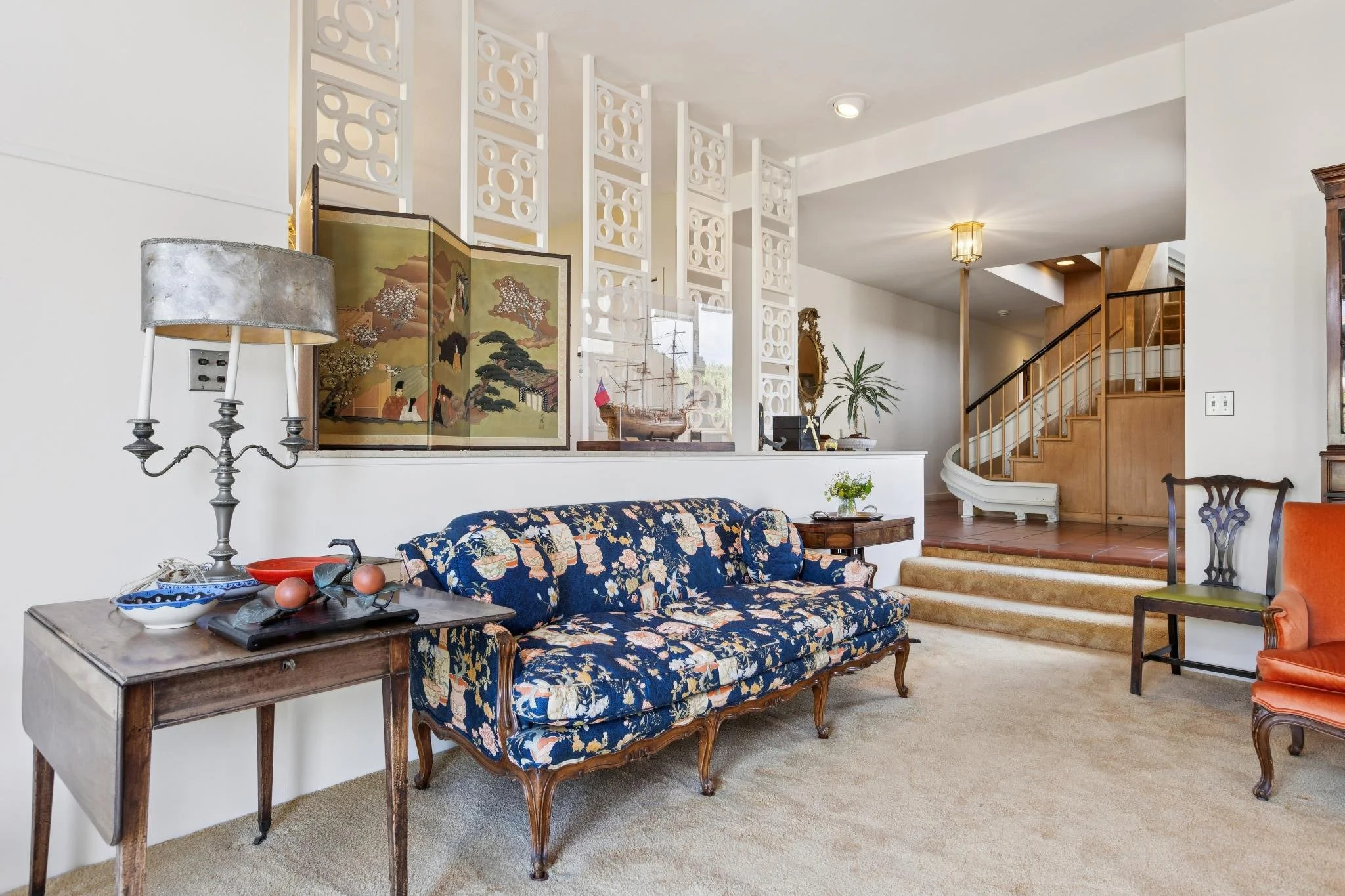 Living room with a floral-patterned sofa, a wooden side table with decorative items, a vintage lamp, and a folding screen with traditional Asian artwork. A staircase with wooden railing is visible in the background, and a high decorative white partit