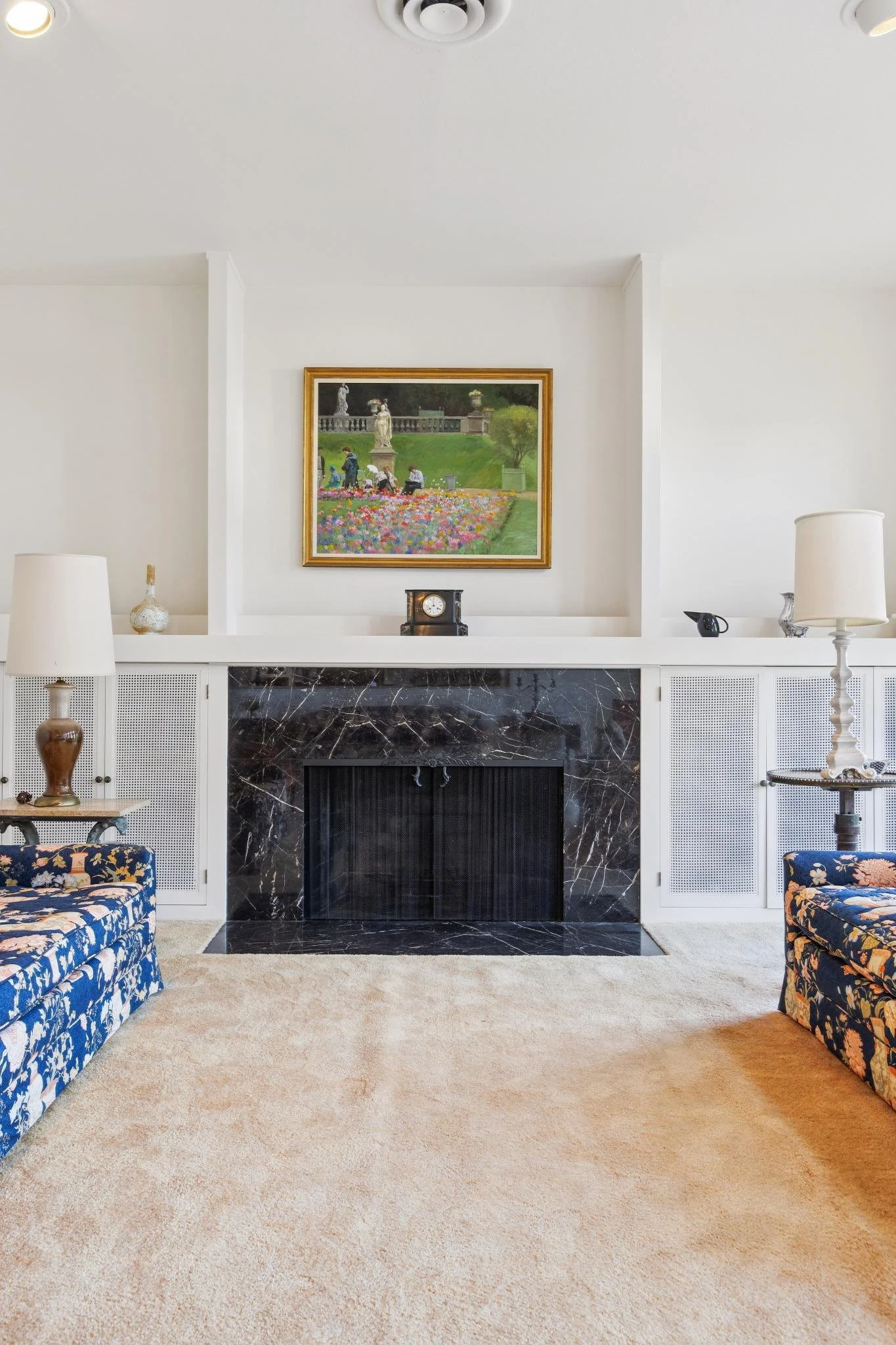 Living room with fireplace, floral-patterned blue armchairs, beige carpet, and mantle with decorative items. Framed painting of a garden scene with sculptures hangs above the fireplace.