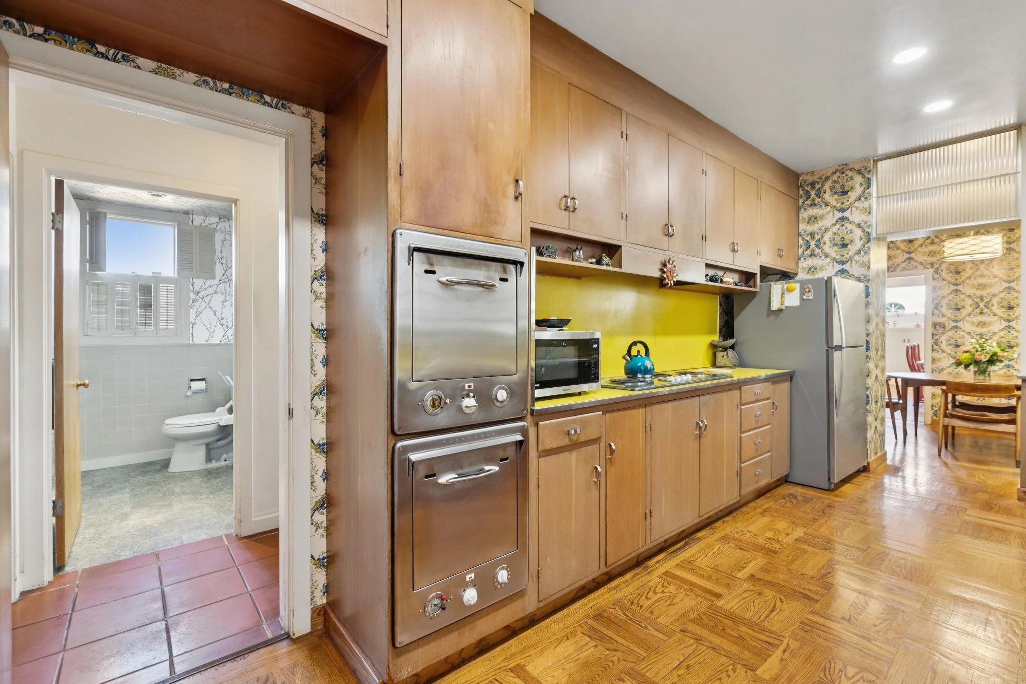 Kitchen with wooden cabinets, yellow backsplash, microwave, refrigerator, and a table with chairs in the background. A doorway leads to a bathroom with a toilet and window.