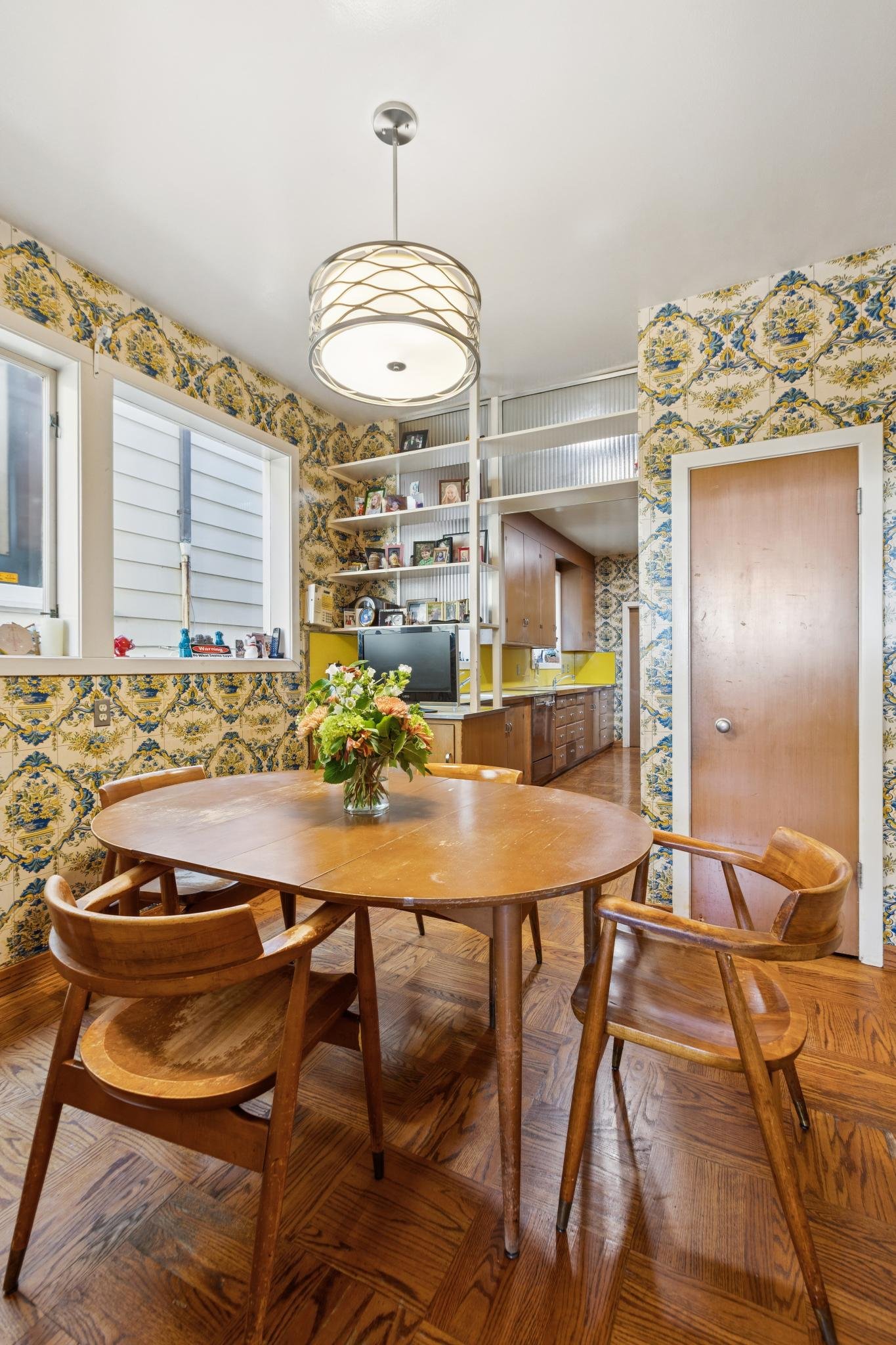 A dining room with a round wooden table surrounded by six matching wooden chairs. The table has a glass vase filled with flowers. The room has vintage-style wallpaper with floral patterns, a ceiling light, a window with blinds, and a corner with shel