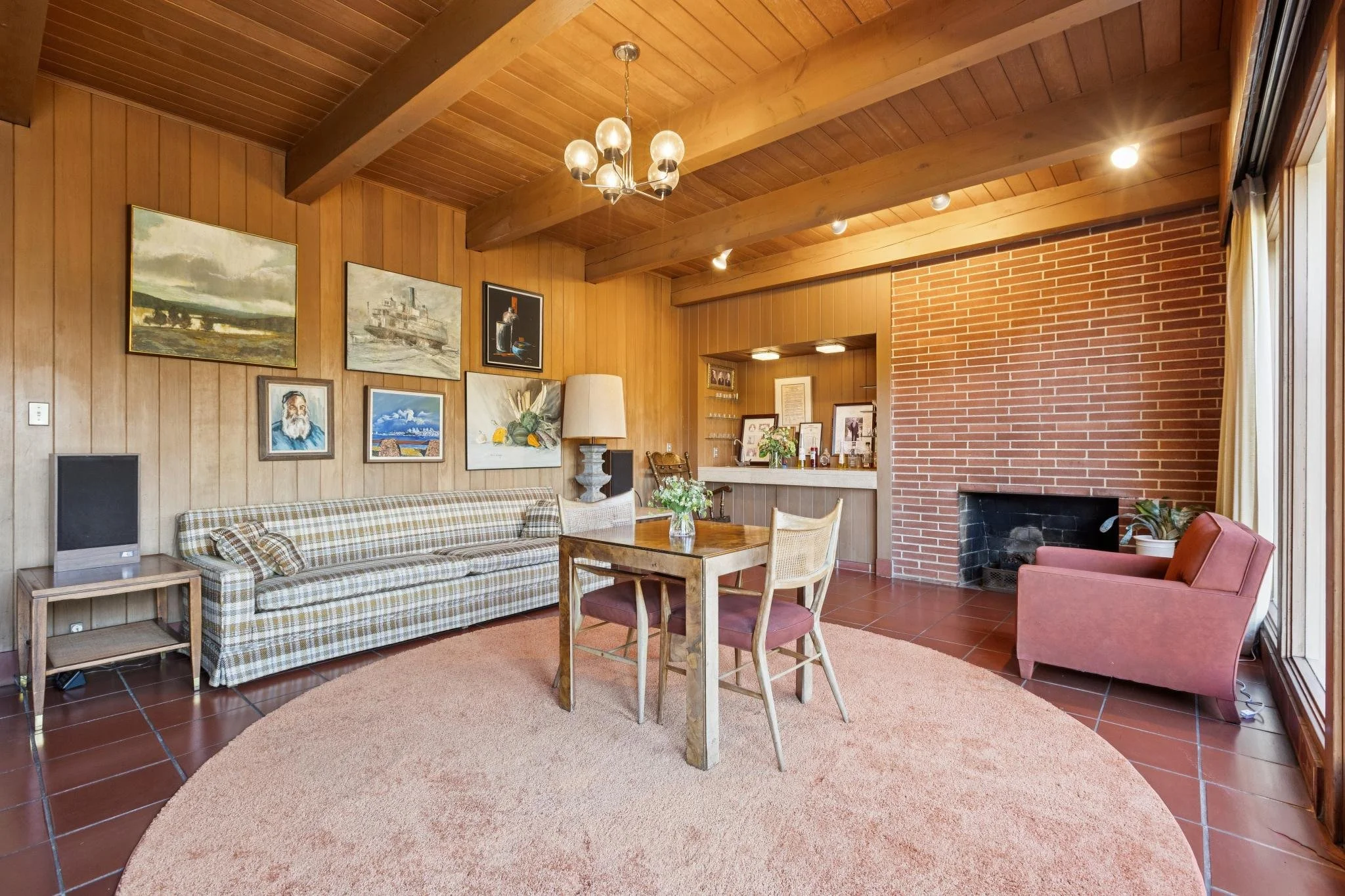 Living room with wood-paneled walls and ceiling, brick fireplace, art paintings on the wall, a checkered sofa, a wooden dining table with chairs, and a pink armchair, with a large window letting in sunlight.