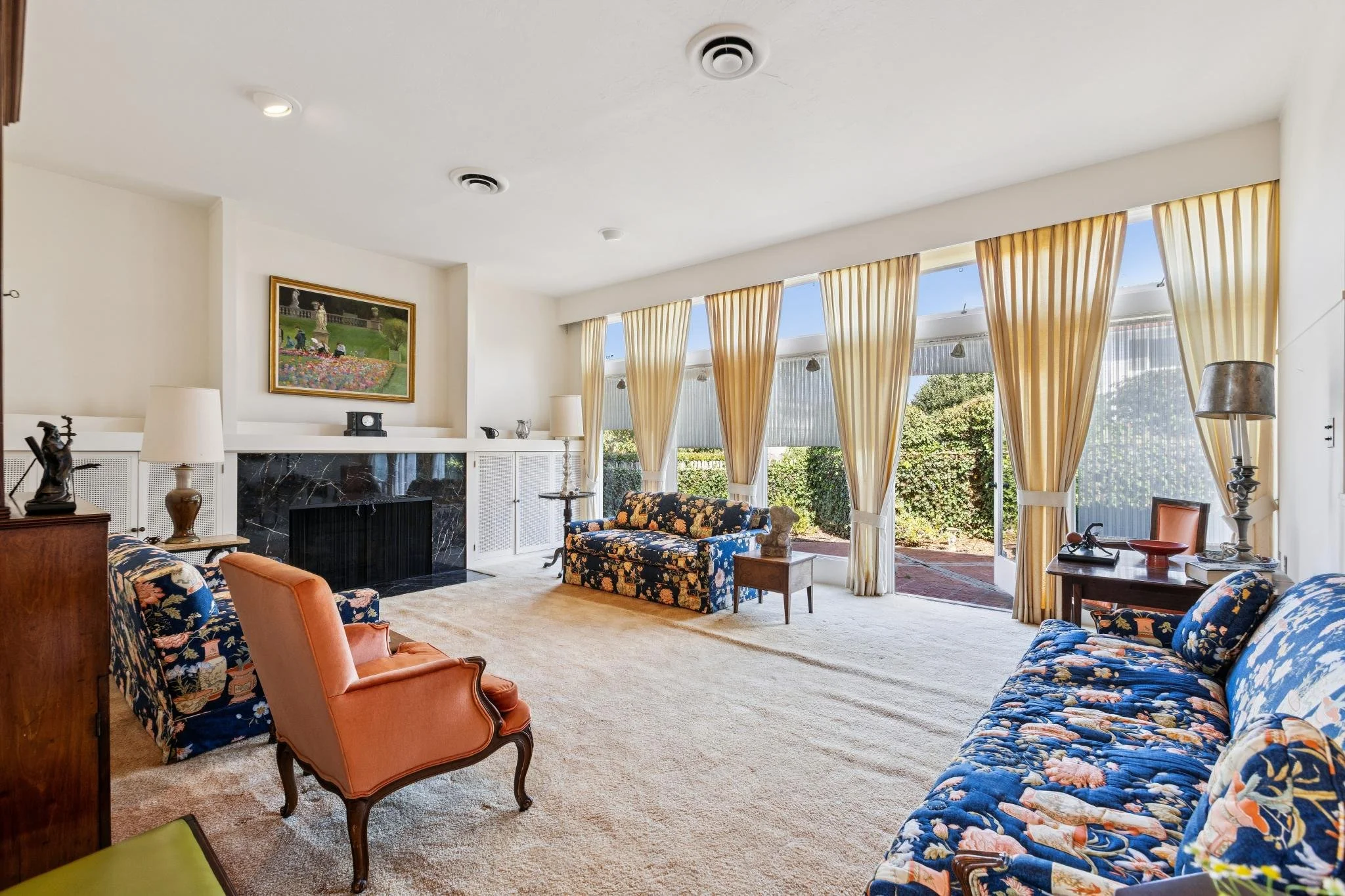 Living room with floral sofas, armchair, fireplace, large window with yellow curtains, artwork above fireplace, decorative items, and outdoor view.