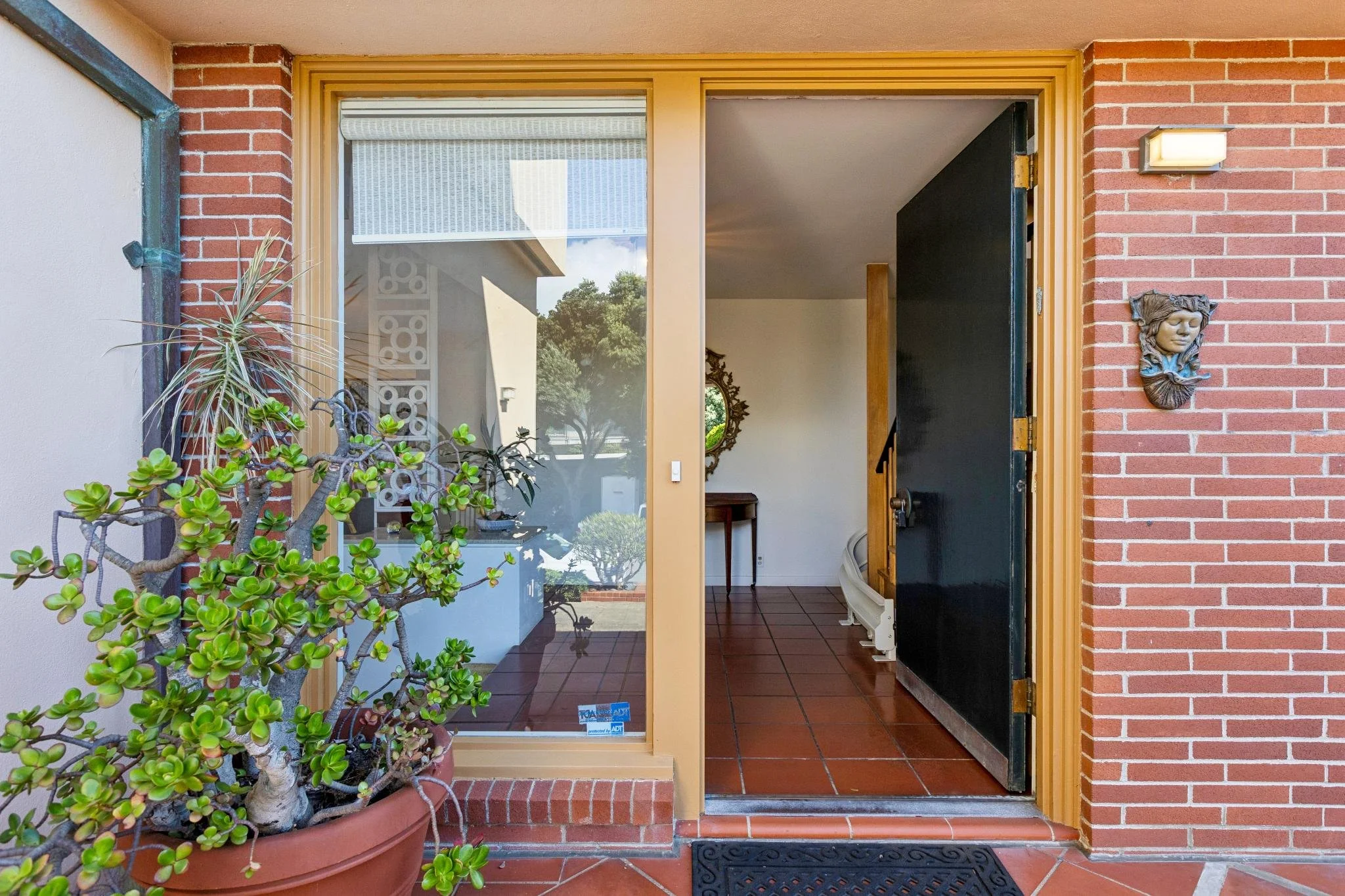 Entrance to a house with a glass door and a black door open. The exterior features red brick and beige trim. There are plants outside the door, including a jade plant in a large pot. Inside, there is a mirror and a table visible in the background.