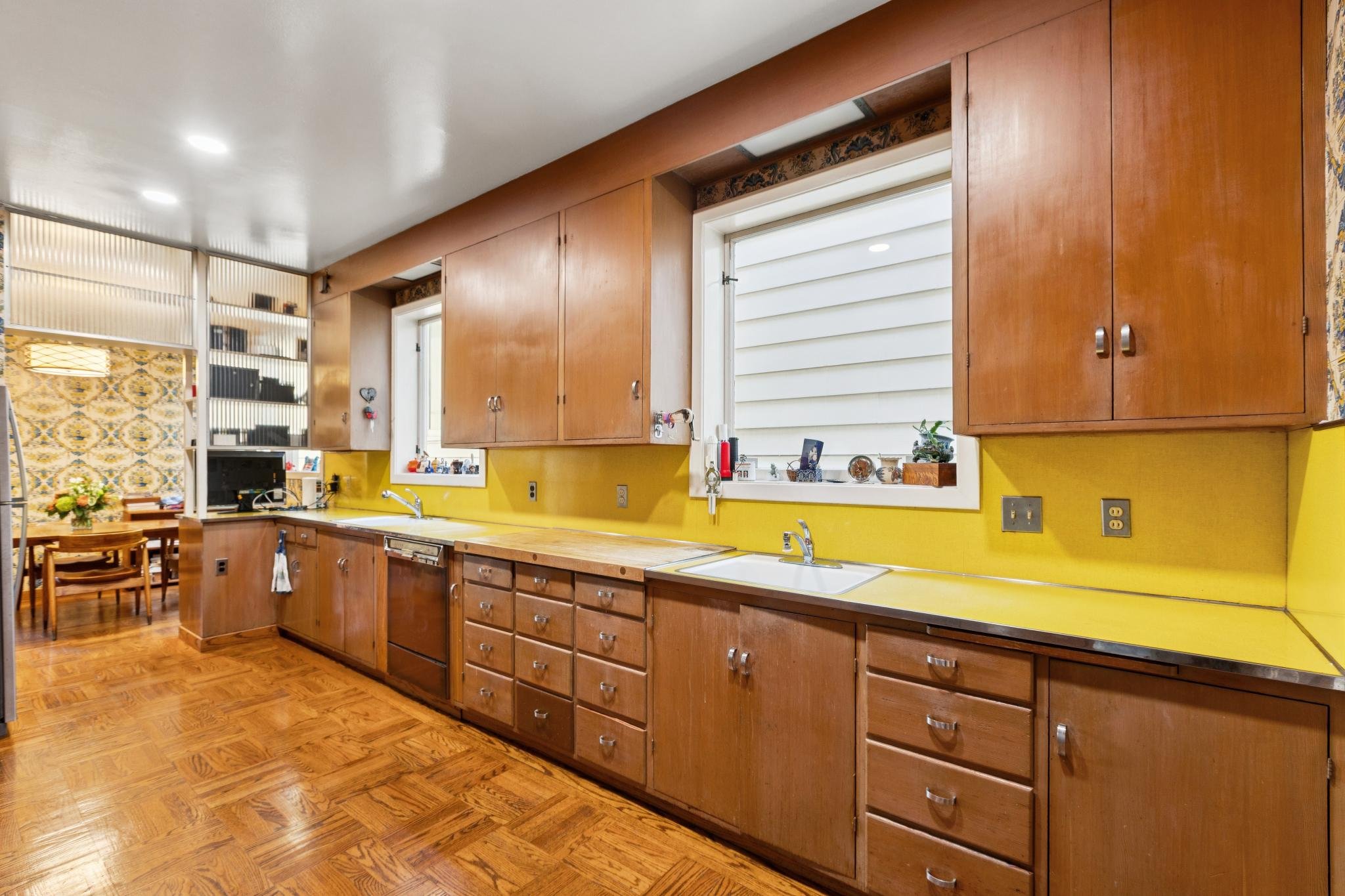 A kitchen with wooden cabinets, yellow walls, a multiple window, and wood flooring. There are some decorations and possible appliances on the countertop.