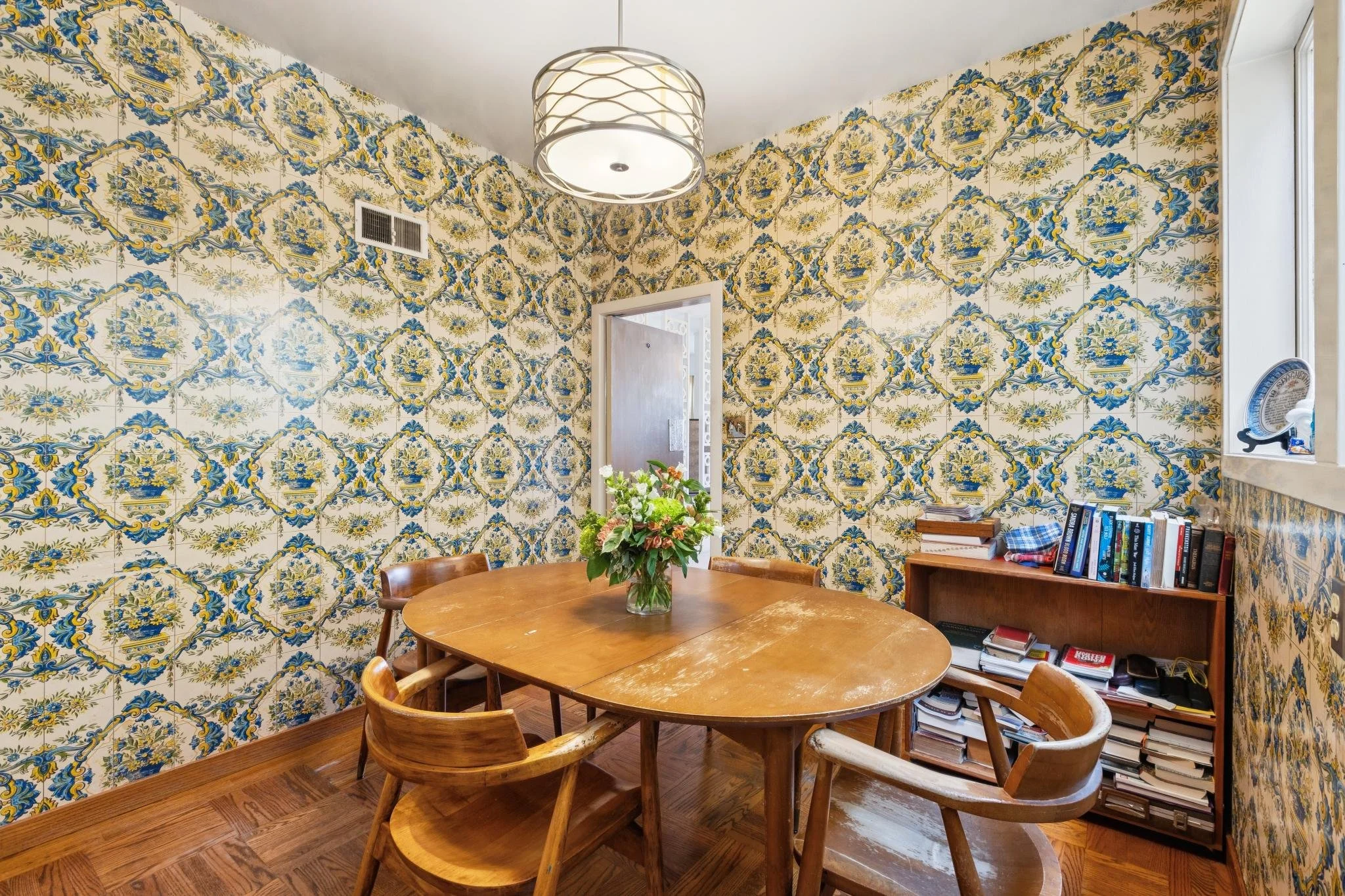 A dining room with vintage floral patterned wallpaper, a wooden table with a vase of flowers, and bookshelves in the corner.