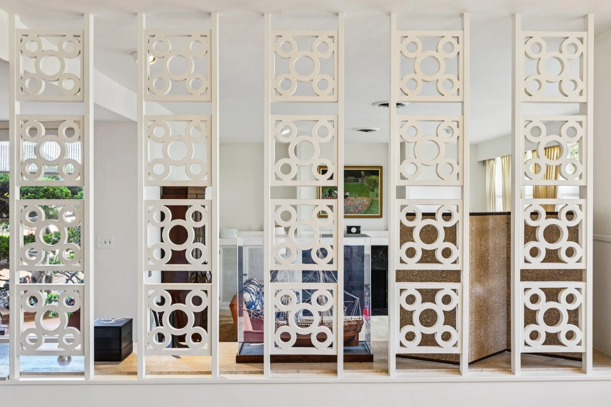 Interior of a house viewed through decorative white partition panels with geometric circular patterns, showing a living area with artwork, furniture, and large windows with curtains.