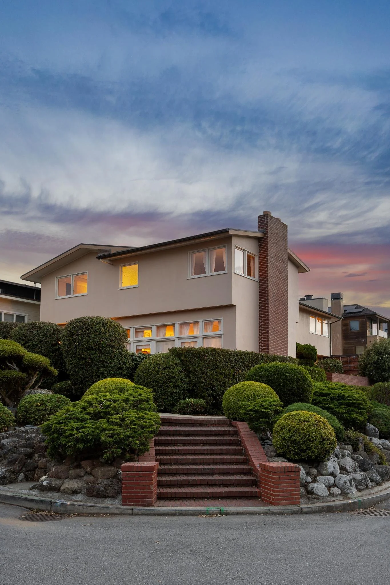A house with a beige exterior and multiple windows, situated on a hill with well-maintained landscaping, including bushes and rocks, at sunset.