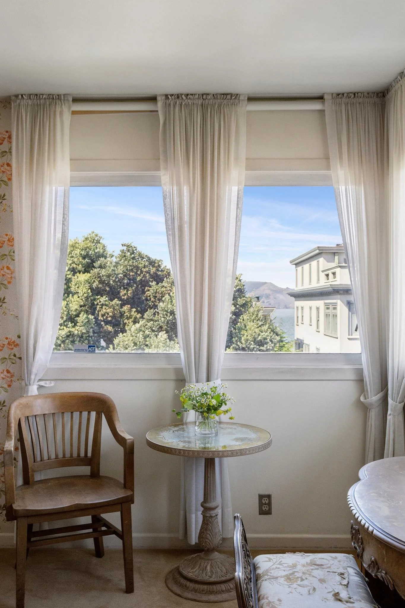 A cozy dining area with a round marble table and a wooden chair. A small flower arrangement on the table. Large windows with white curtains revealing a view of trees, a building, and mountains in the distance.