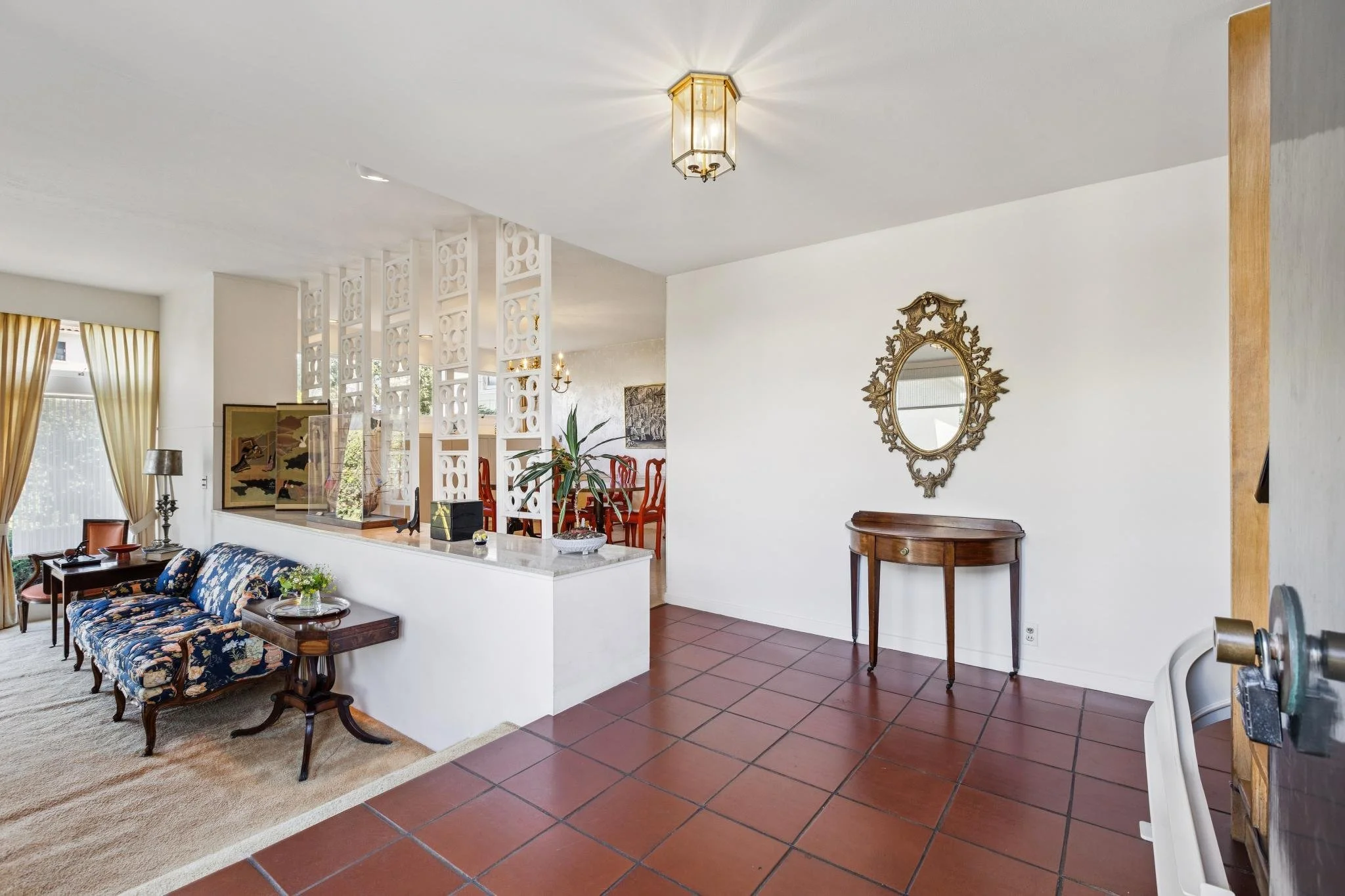 Living room with floral sofa, side tables, lamp, and a decorative mirror on white wall, leading to dining area with red chairs and artwork.