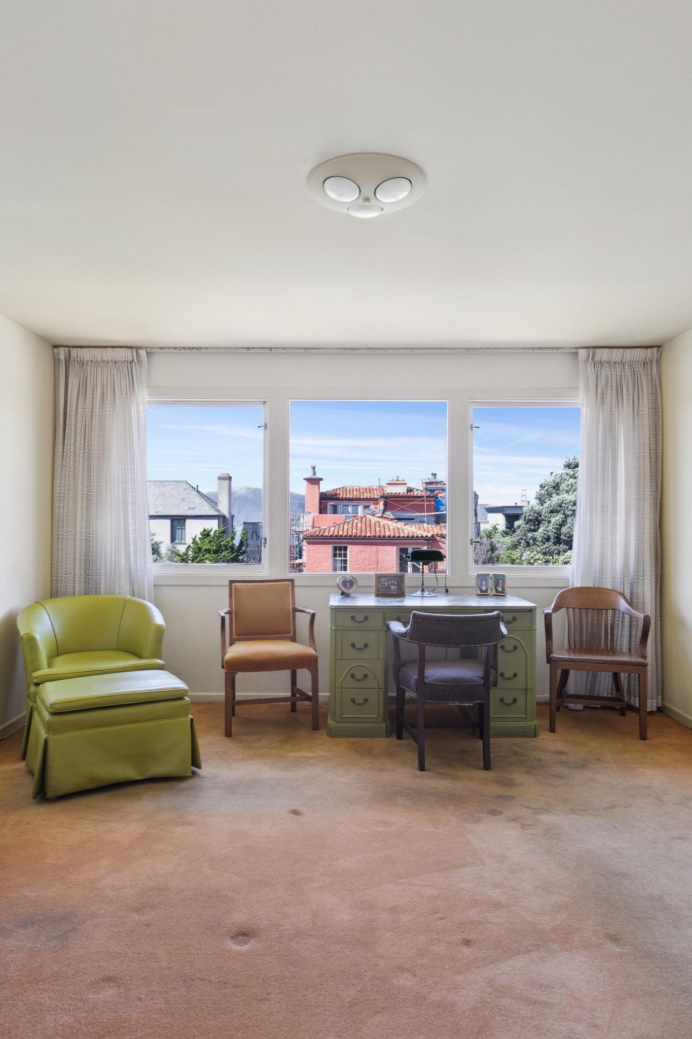 A spacious room with a large window showing rooftops and a blue sky. The room has beige carpet, cream-colored walls, and a variety of chairs including a lime green armchair, a wooden chair, two chairs at a green desk, and a dark gray chair. There are