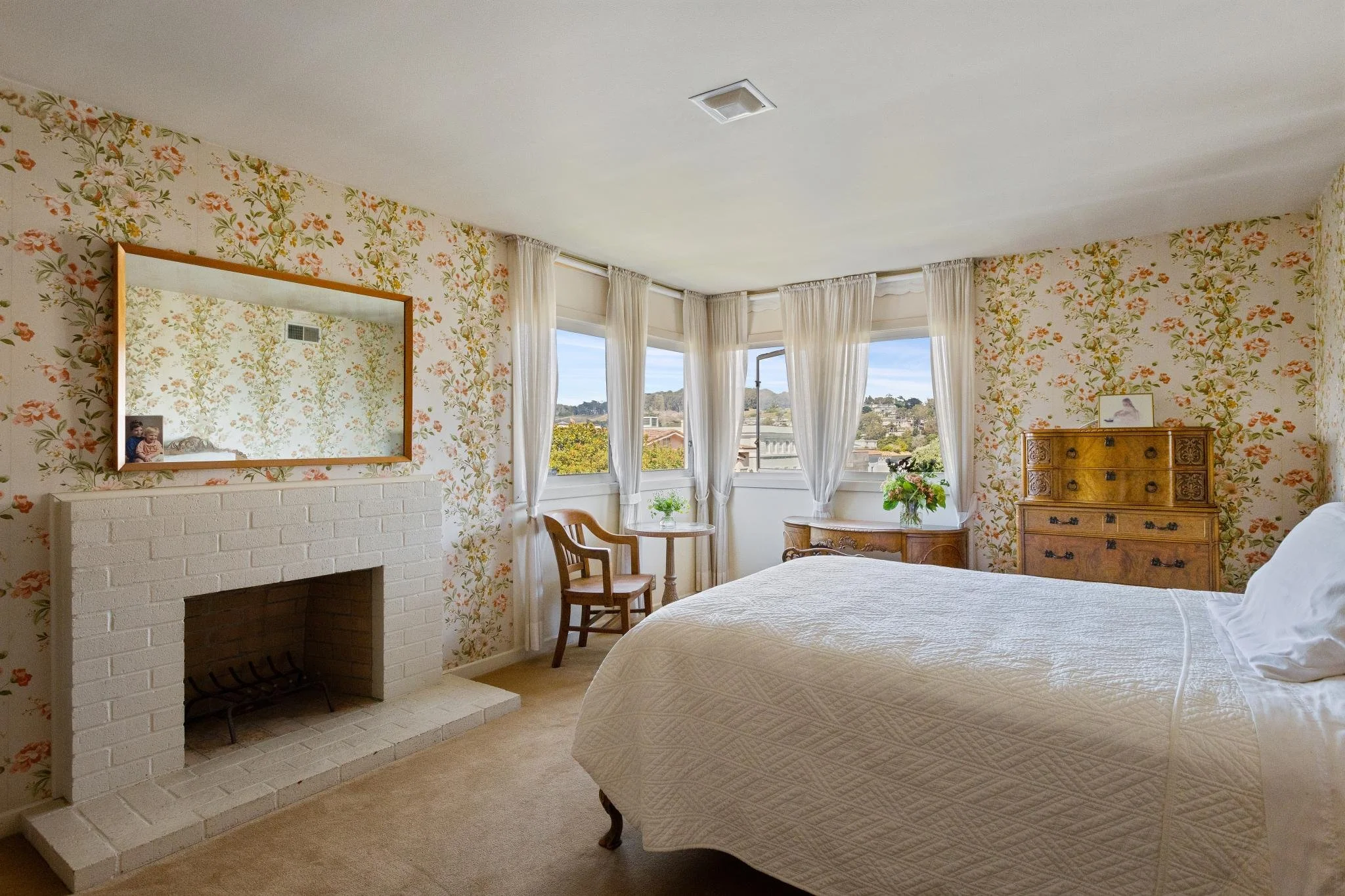 A bedroom with floral wallpaper, a white brick fireplace, a mirror, and a window with curtains letting in natural light.