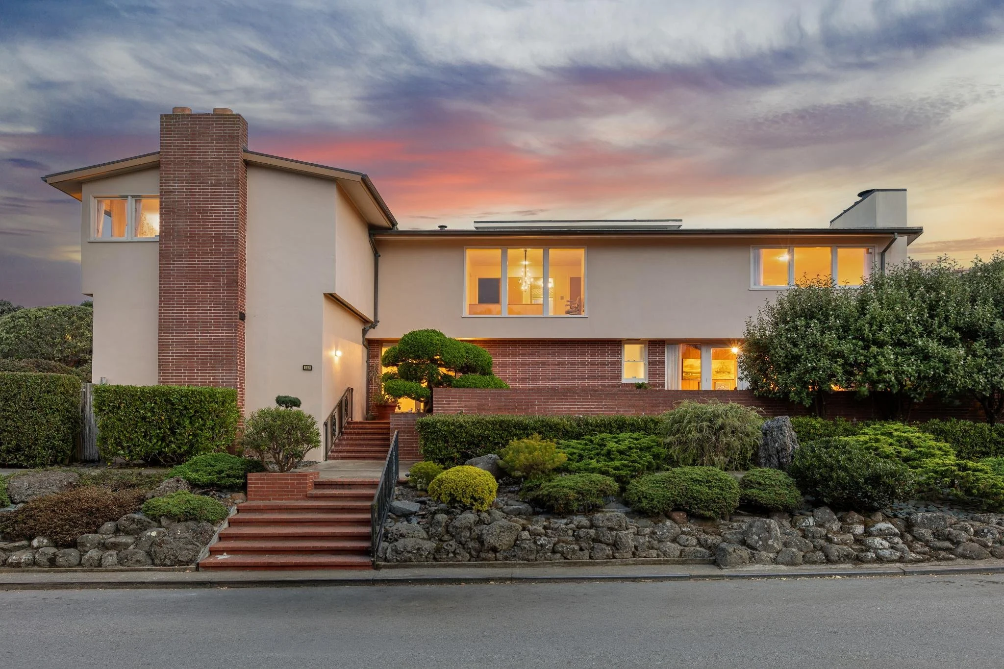 A modern two-story house with beige and brick exterior, large illuminated windows, stone-lined front yard, and landscaped garden, during sunset with a colorful sky.