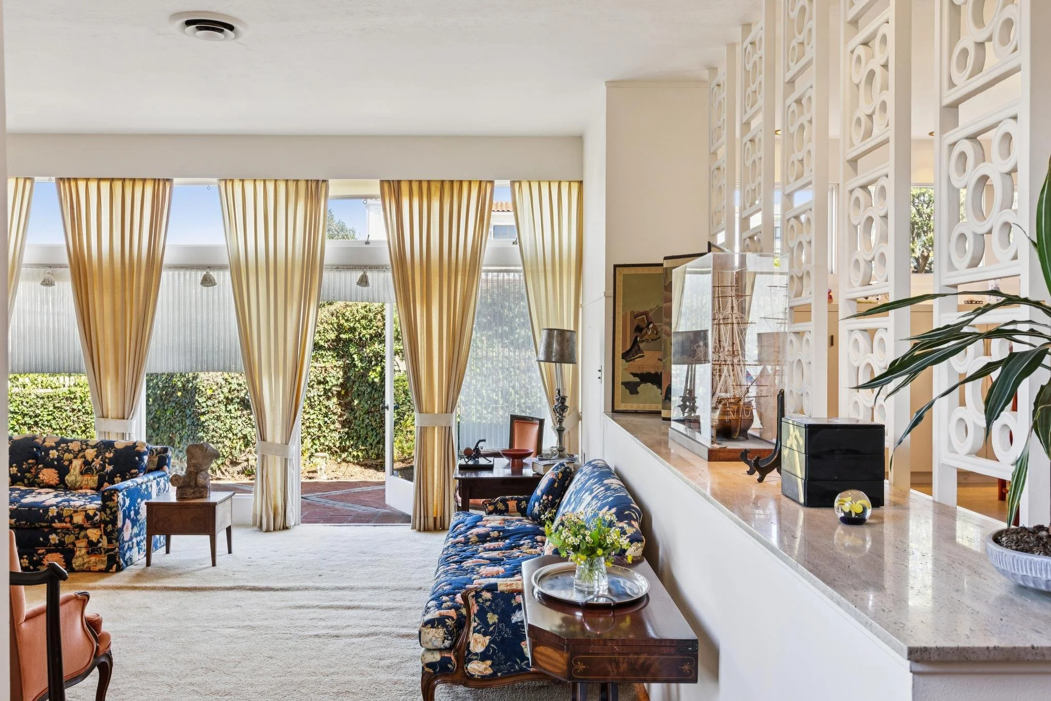 Spacious living room with large windows, yellow curtains, floral-patterned sofa, assorted chairs, and decorative items on a countertop and side tables.