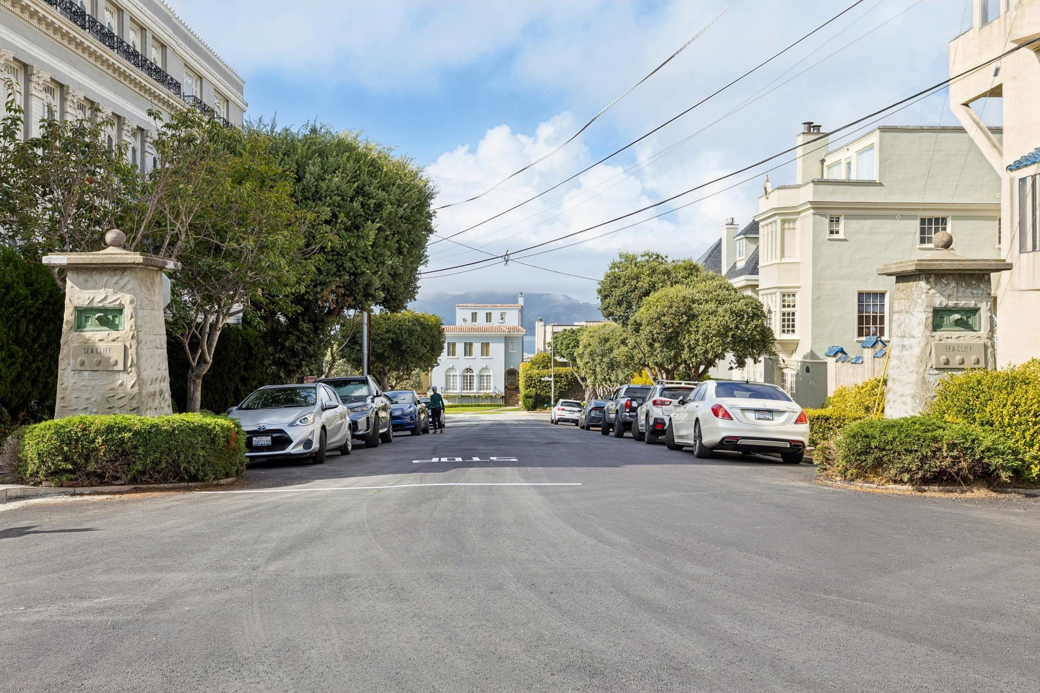 Sea Cliff Neighborhood in San Francisco. Residential street with parked cars, large trees, and multi-story homes with ornate architecture under a partly cloudy sky.
