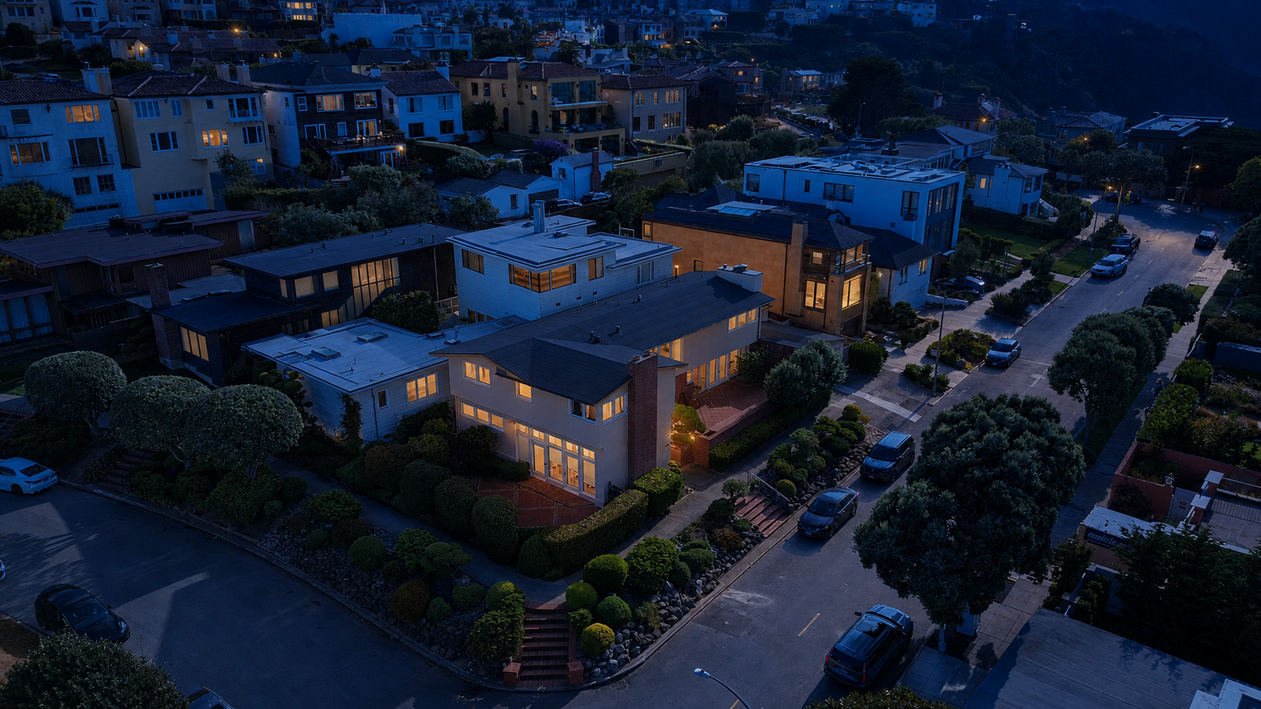 Aerial view of a residential neighborhood, Sea Cliff in San Francisco, during dusk, showing modern houses with lit windows, trees, parked cars, and a street.
