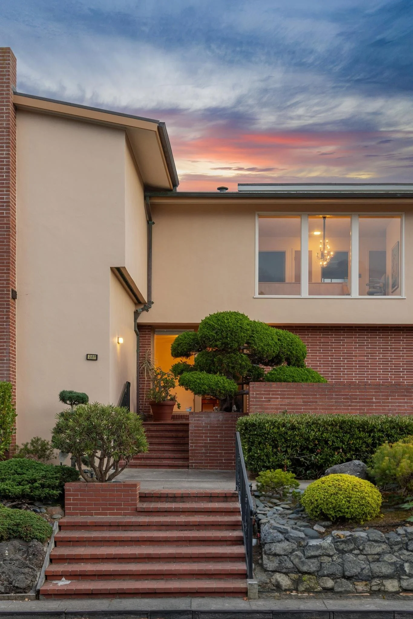 Exterior of a modern house with a brick front, large window, and well-maintained garden with bushes and small trees, under a colorful sunset sky.