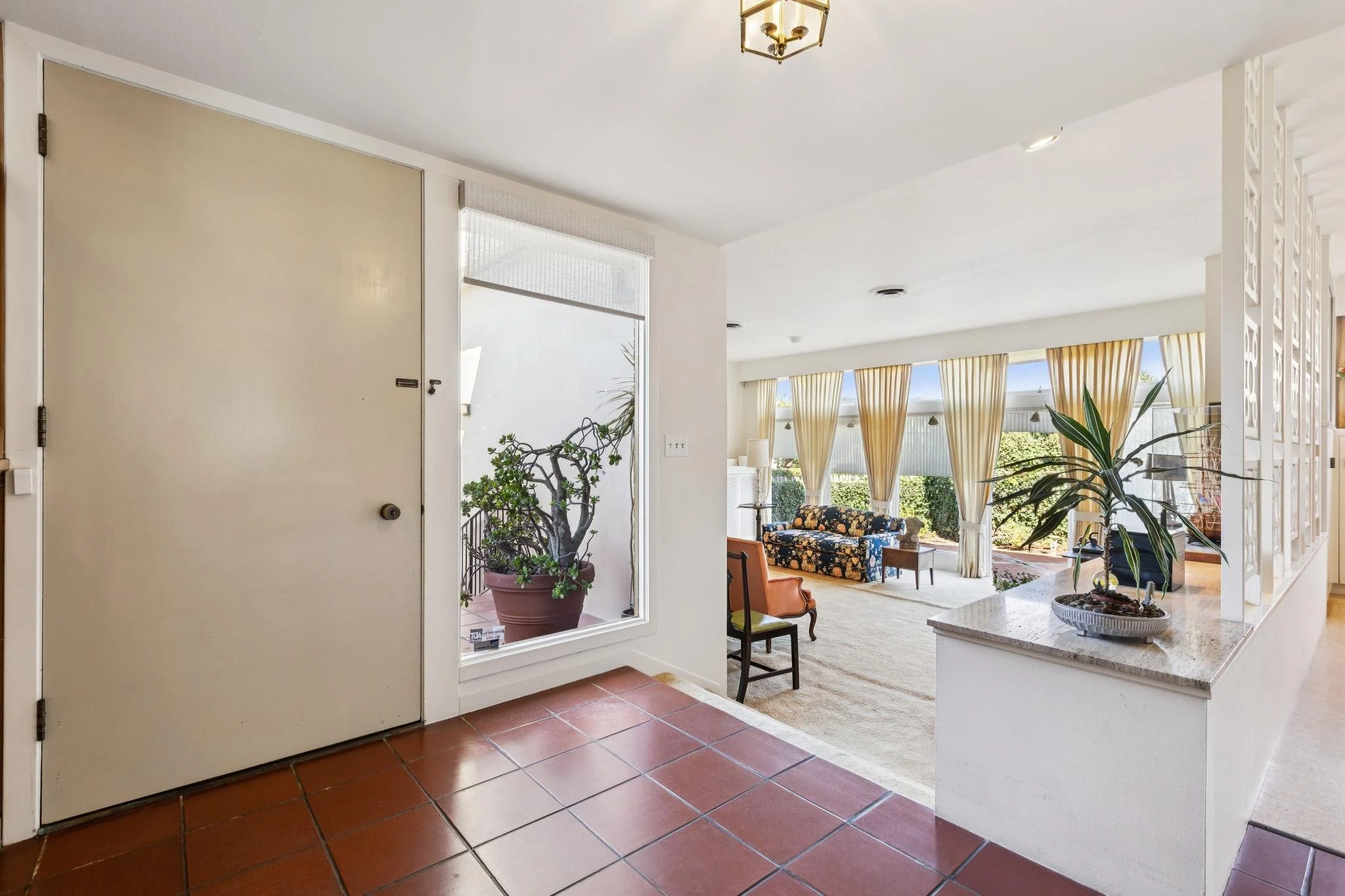 Interior view of a living room seen through the open front door. It features large windows with yellow curtains, a floral sofa, potted plants, and a beige carpet on a hardwood floor.