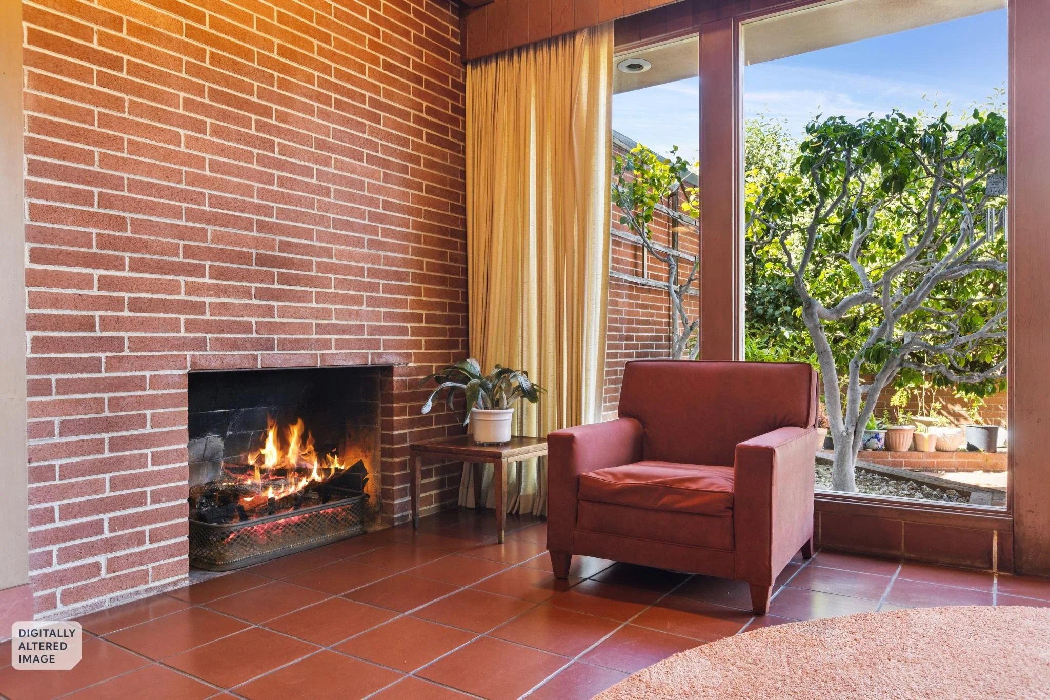 Living room with brick fireplace, red armchair, small wooden side table with a potted plant, large glass window showing green trees outside, and yellow curtains.