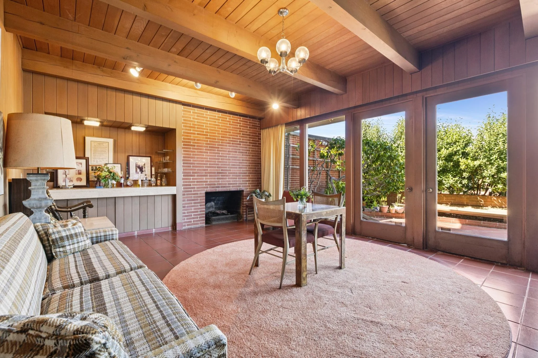 A living room with wooden paneling on the walls and ceiling, a brick fireplace, large windows facing outside with trees, a sofa with plaid cushions, a table with chairs, a standing lamp, and decorative items on the mantle.