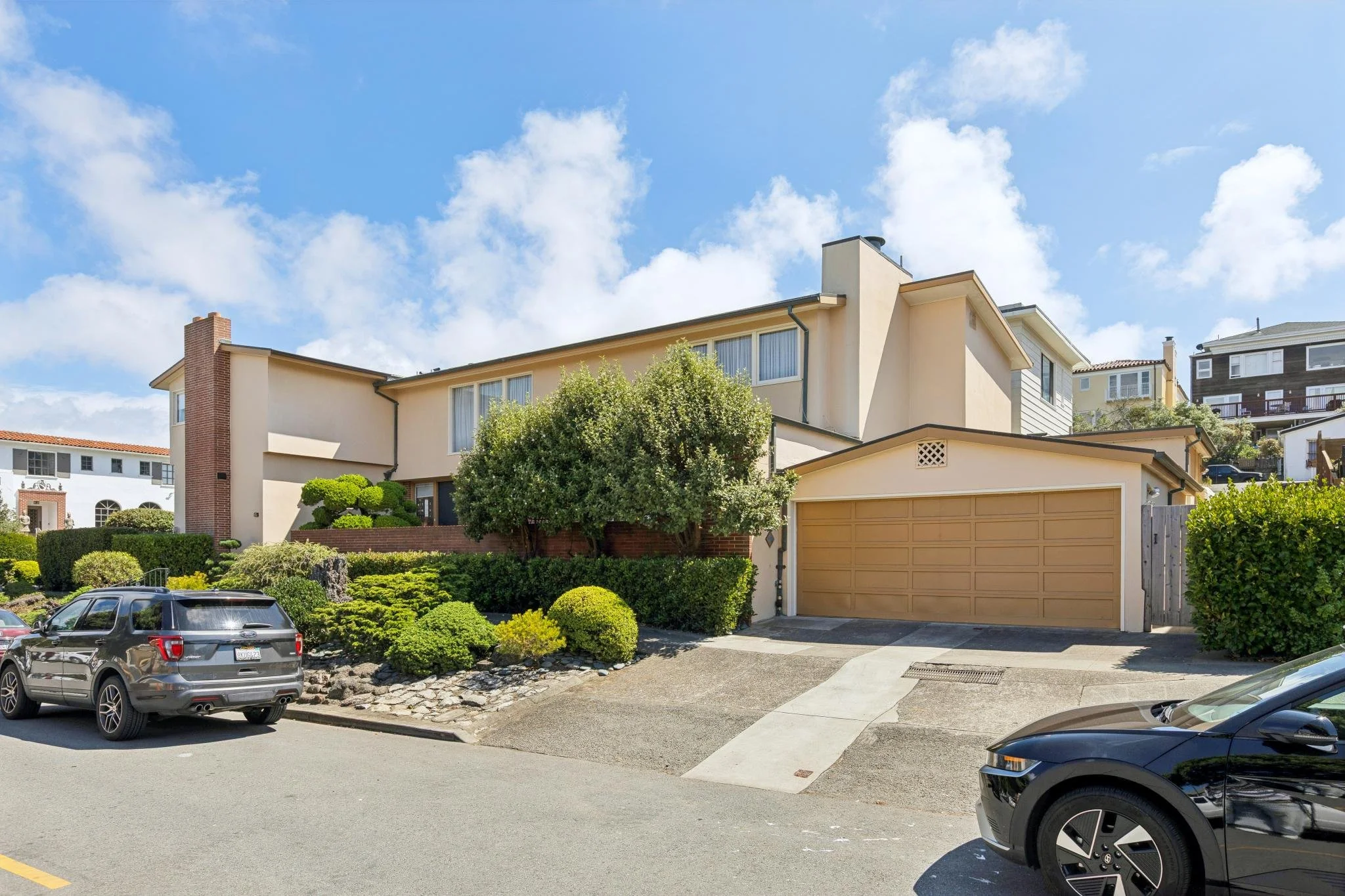 A residential street scene with parked cars in front of a beige house with multiple stories and large windows, surrounded by green bushes and trees, under a partly cloudy blue sky.