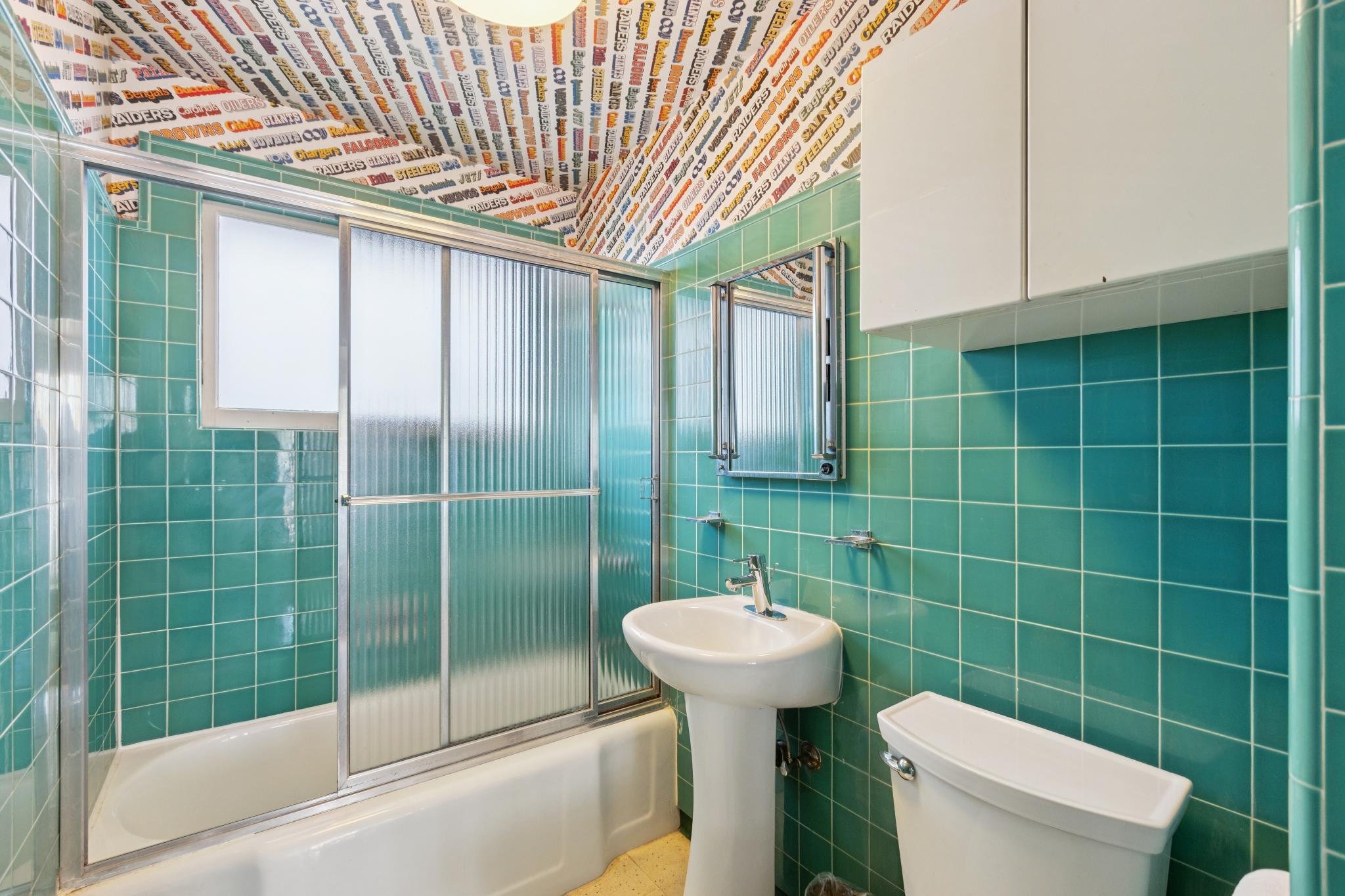 A bathroom with green tiled walls and a frosted glass shower door. Inside the bathroom, there is a white bathtub, a white toilet, a white pedestal sink, and a medicine cabinet. The ceiling has wallpaper with sports team names and logos.