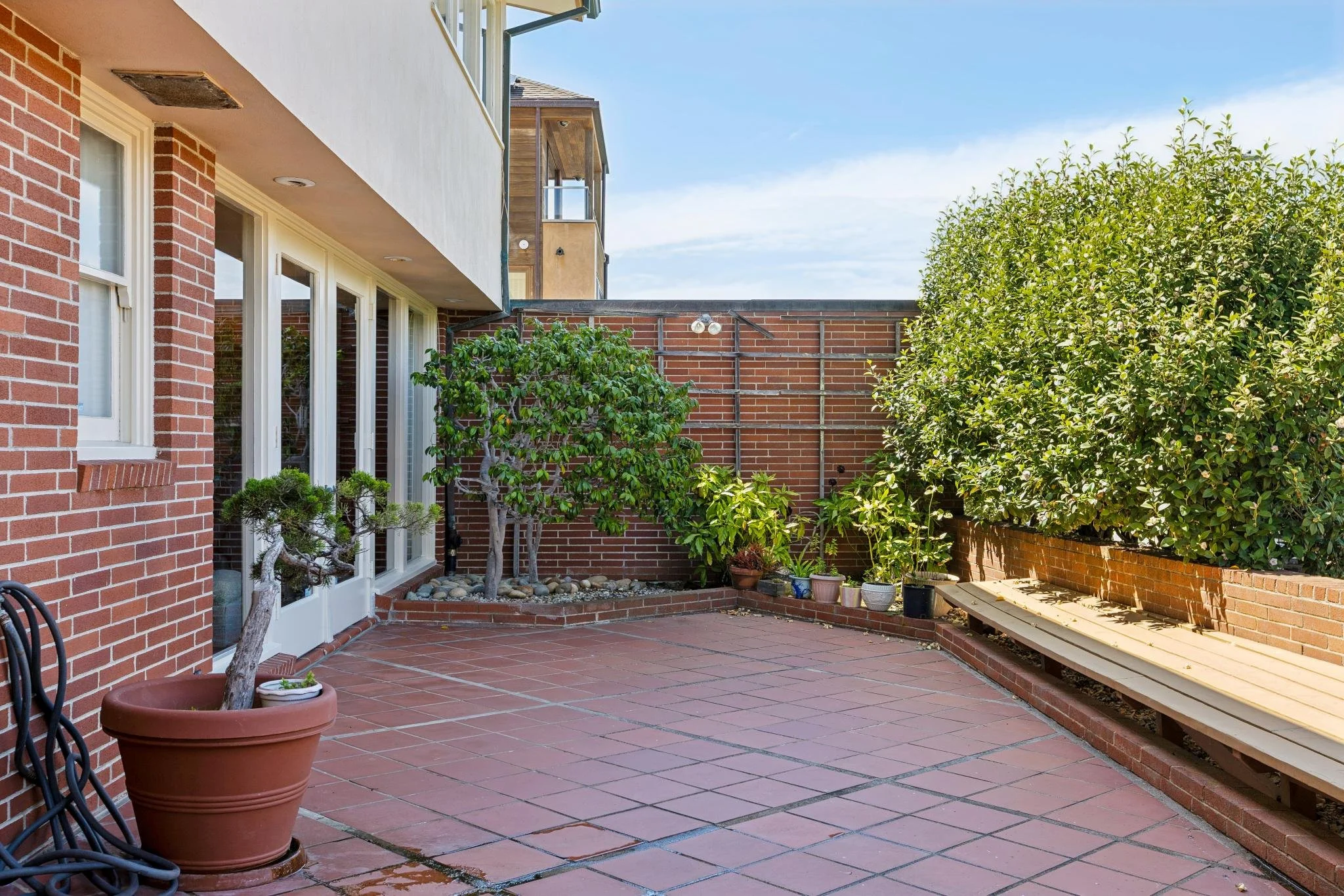 A backyard patio area with brick walls, potted plants, a small tree, and a brick fence. The patio has red tiles and a wooden bench along the brick wall. There are several small potted plants on the ground and a garden hose coiled on the left.