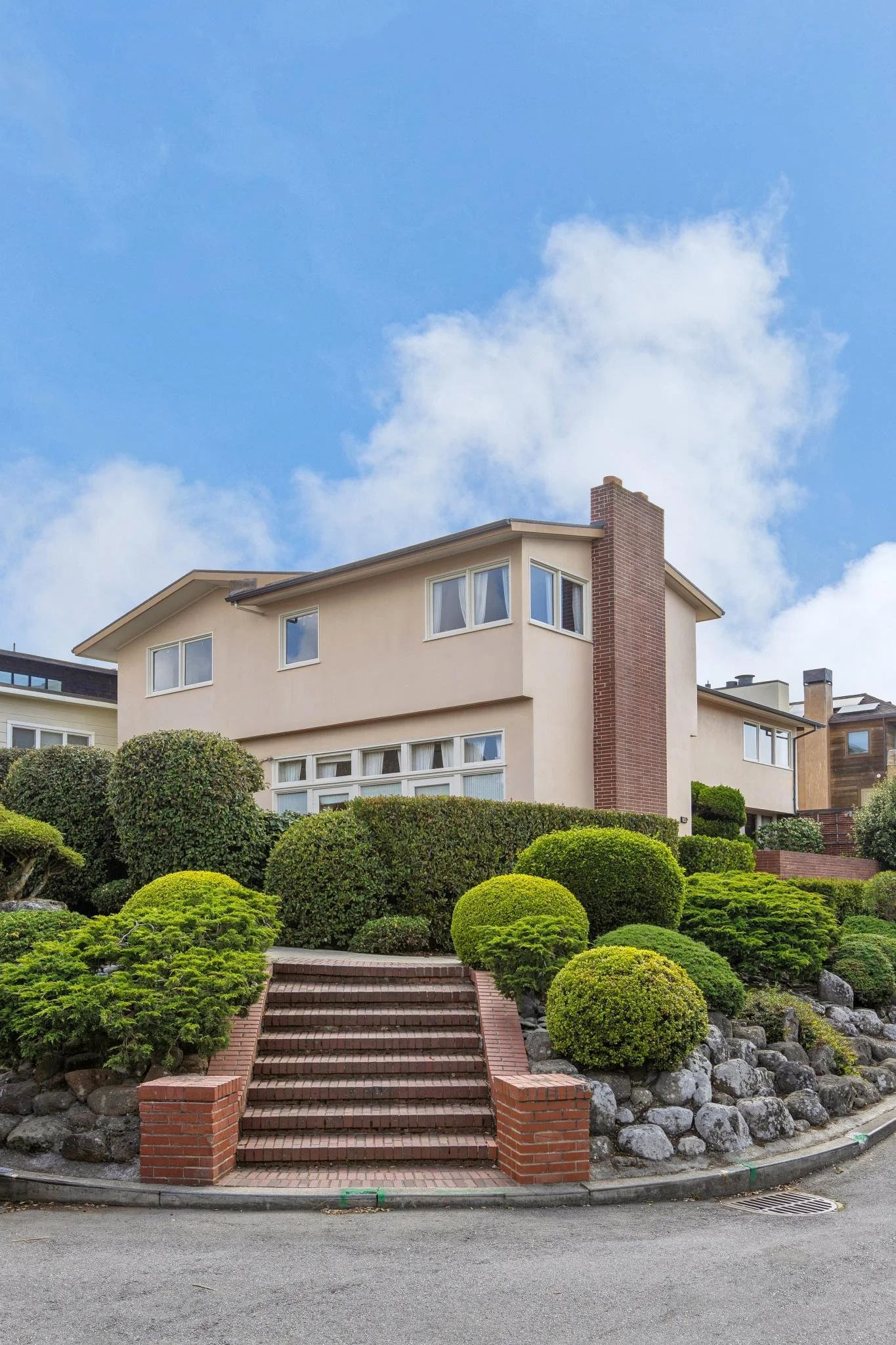 Front view of a modern multi-story house with a landscaped garden, brick stairs leading to the entrance, and a blue sky with clouds.
