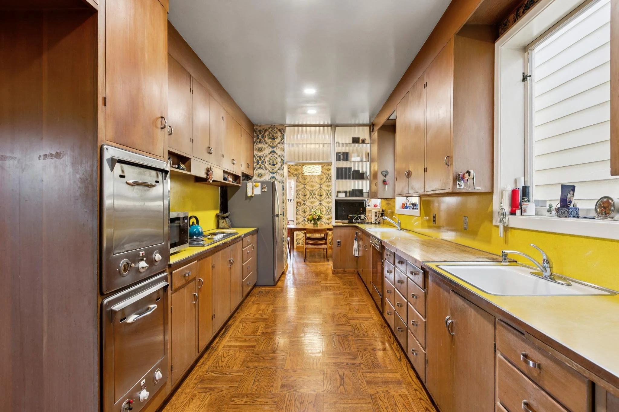 A vintage kitchen with wooden cabinets, yellow countertops, a stainless steel oven, a fridge, and a window with white siding outside. There's a small dining table with chairs in the background.
