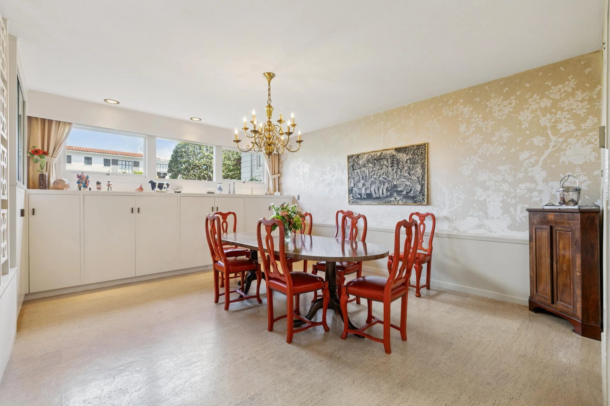 A dining room with a wooden oval table surrounded by nine red wooden chairs, a chandelier hanging from the ceiling, a large window with curtains, and a framed artwork on the wall.