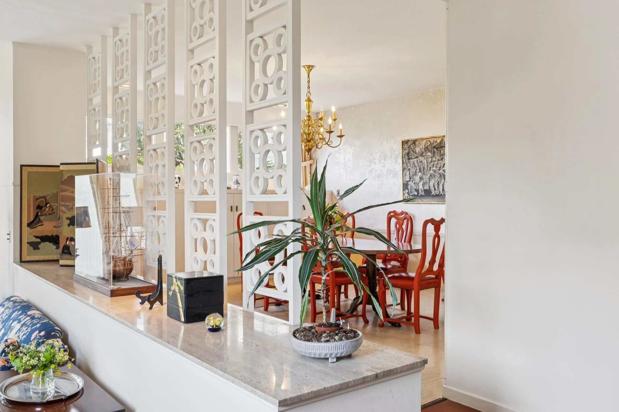 A dining room with red chairs around a wooden table, a chandelier hanging from the ceiling, and a decorative screen dividing the space. There are multiple framed artworks on the wall and large windows letting in natural light. Various plants and deco