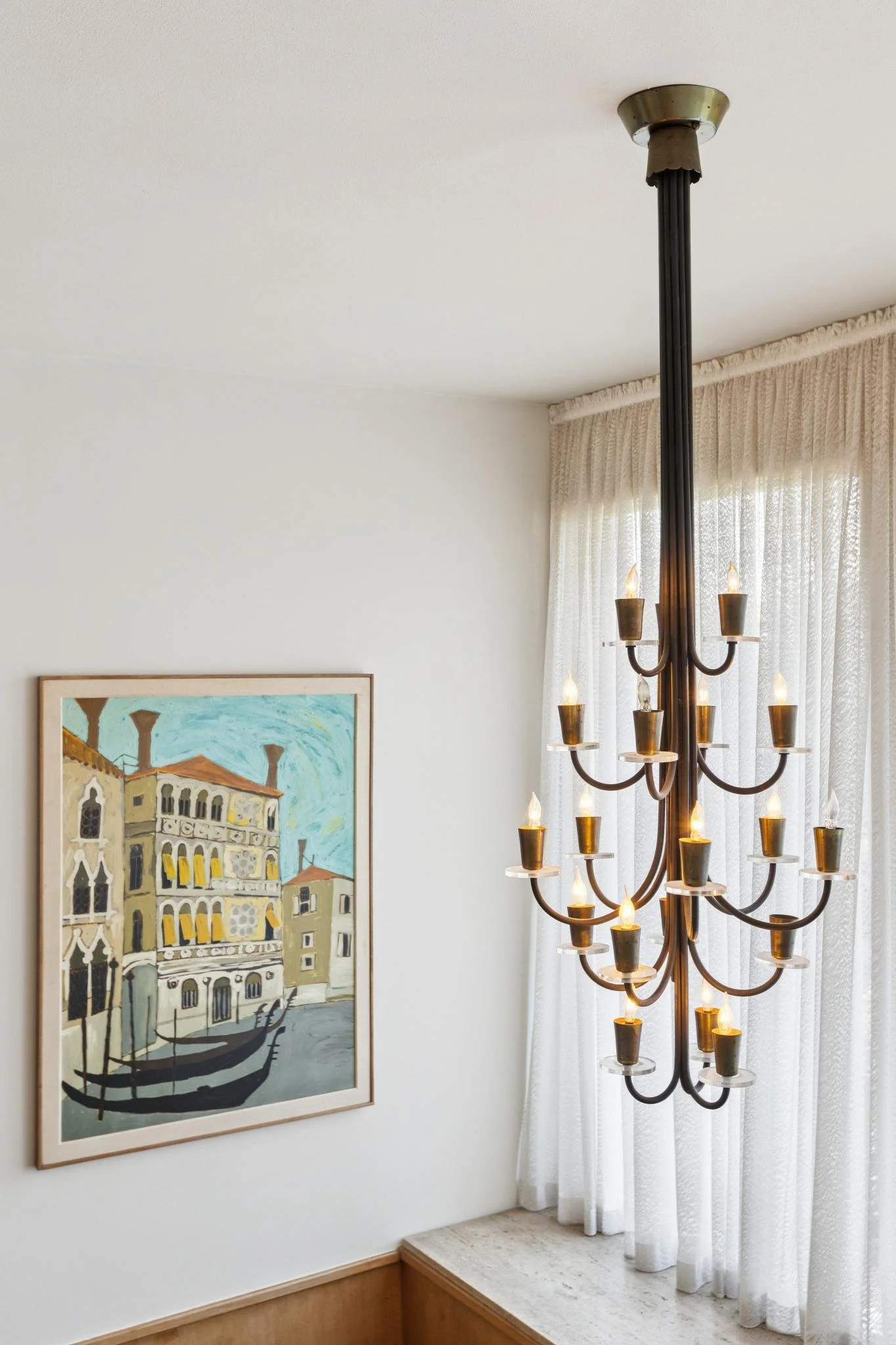 Living room with a framed painting of buildings on the wall, a multi-armed chandelier, and window curtains letting in natural light.