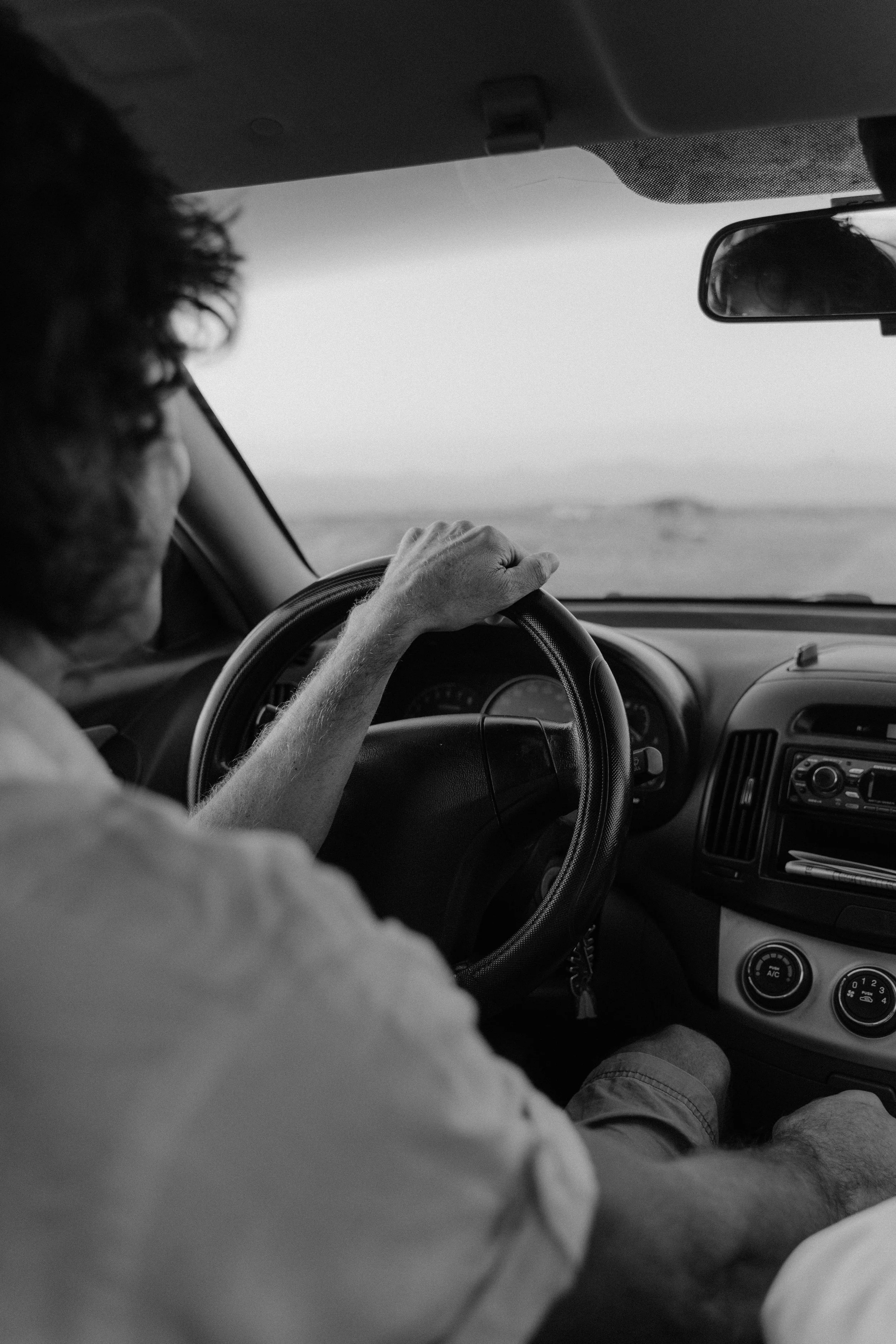 Vehículo en movimiento en un camino desértico, vista desde el interior, con el conductor de cabello rizado y manos sobre el volante, en un día despejado.