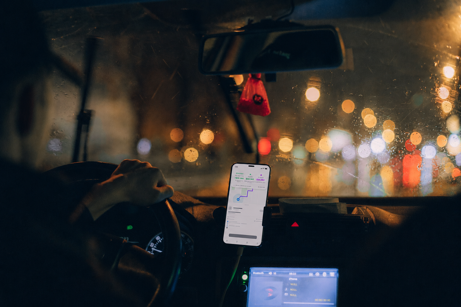 Vista del interior de un coche en la noche, con la lluvia en las ventanas y luces borrosas del exterior, mostrando una mano en el volante y un teléfono móvil con una app de navegación en el soporte del coche.