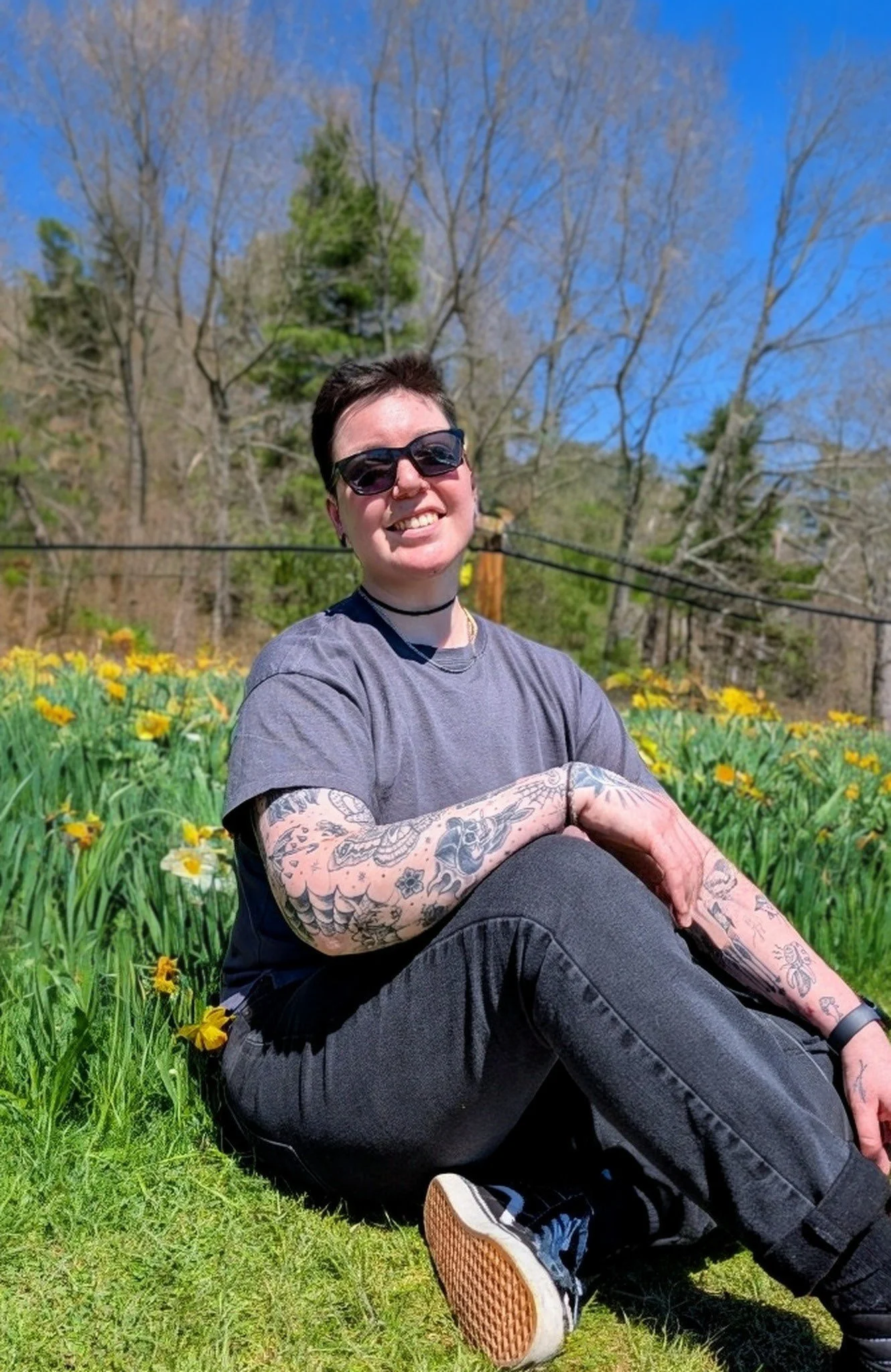 A person with short dark hair, sunglasses, tattoos on arms, wearing a choker, grey t-shirt, black pants, and sneakers, sitting outdoors on grass with yellow flowers, trees, and a blue sky in the background, smiling.