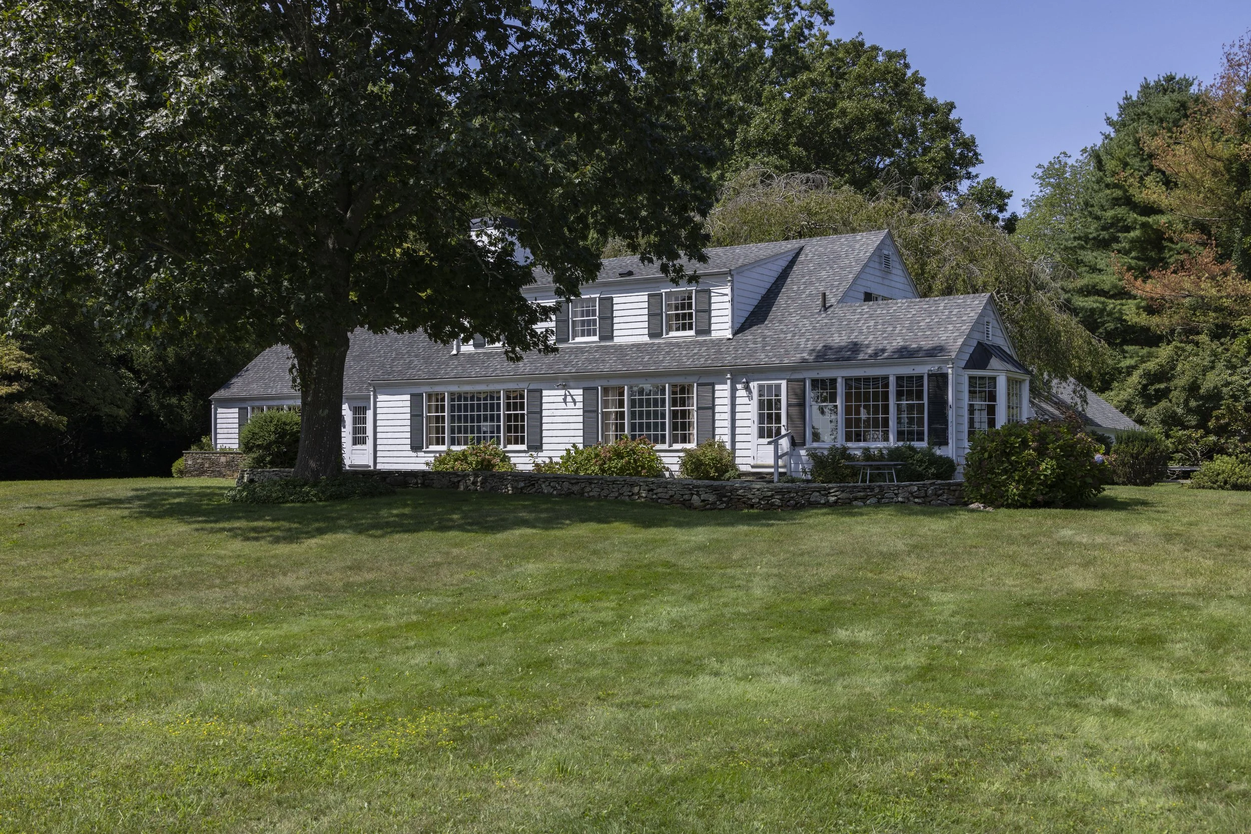 A white house with black shutters and multiple windows, surrounded by lush green trees and an open grassy lawn, under a clear blue sky.