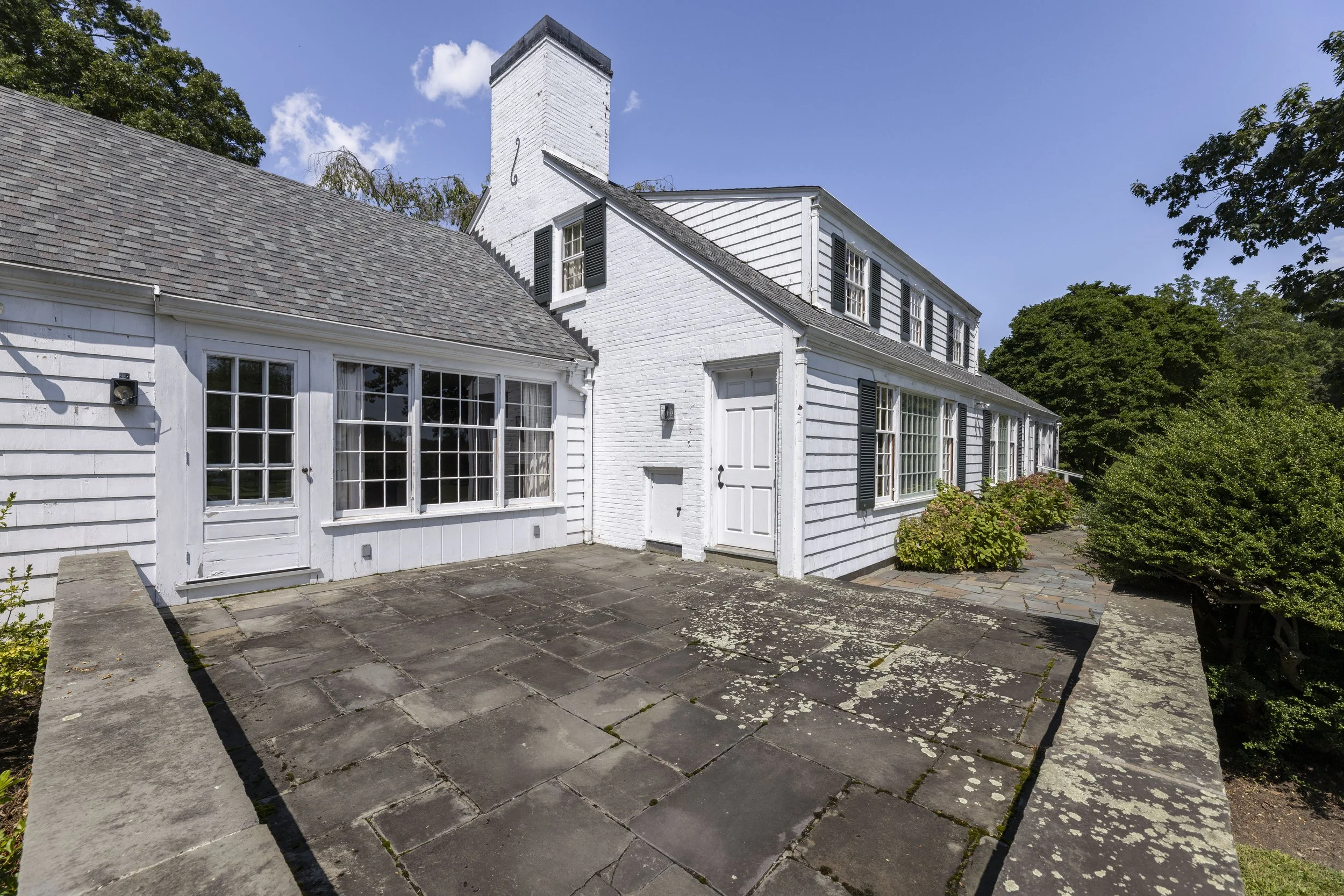 White house with black shutters, brick chimney, large front windows, gray shingle roof, paved patio, surrounded by green trees and bushes, under a blue sky with a few clouds.