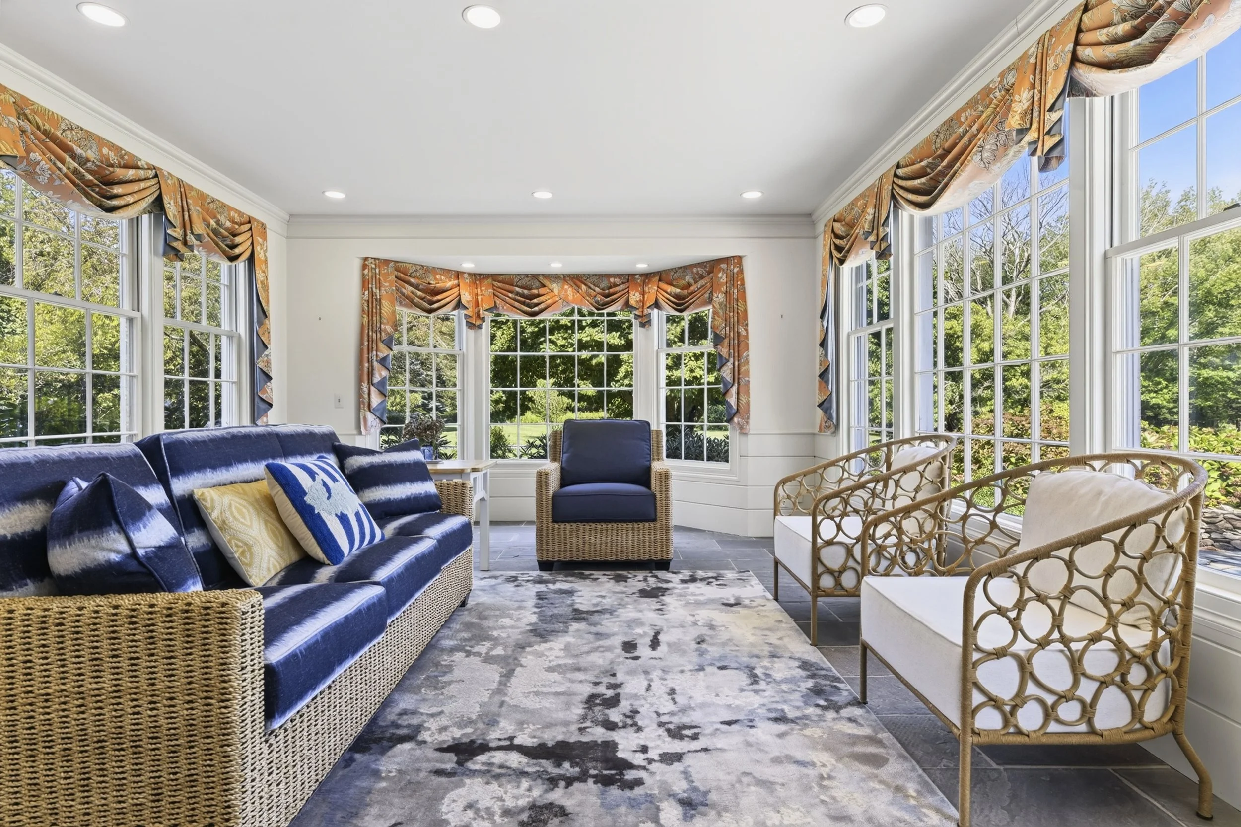 Bright sunlit living room with large windows and colorful curtains, decorated with a blue velvet sofa, wicker chairs, and various decorative pillows, overlooking a lush green garden.