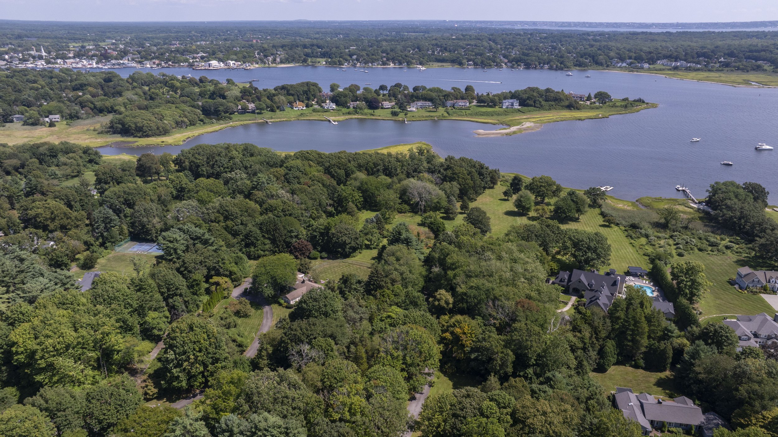 Aerial view of a large body of water with boats, surrounded by green trees and residential houses with lawns, and a distant shoreline with more buildings.