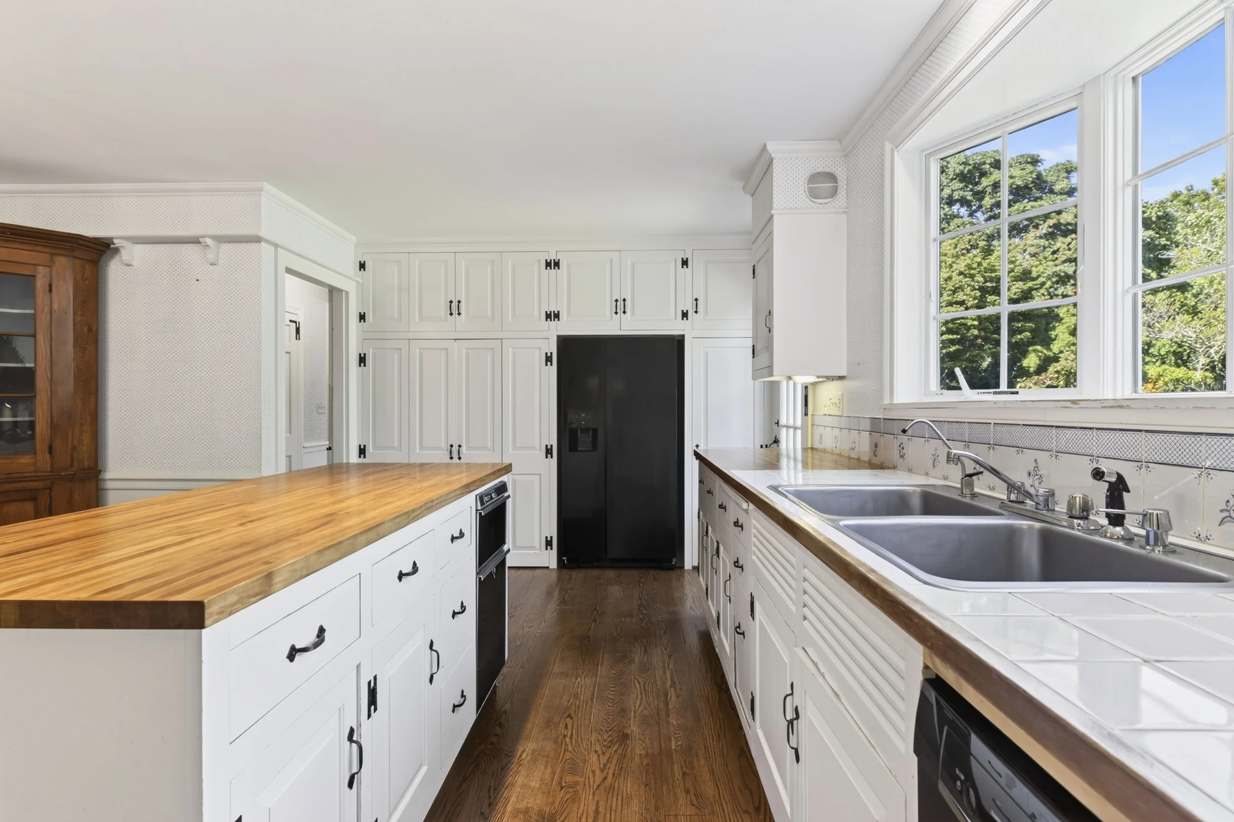 A bright kitchen with large windows showing green trees outside. The kitchen has white cabinets with black handles, a wooden countertop, and a stainless steel sink.