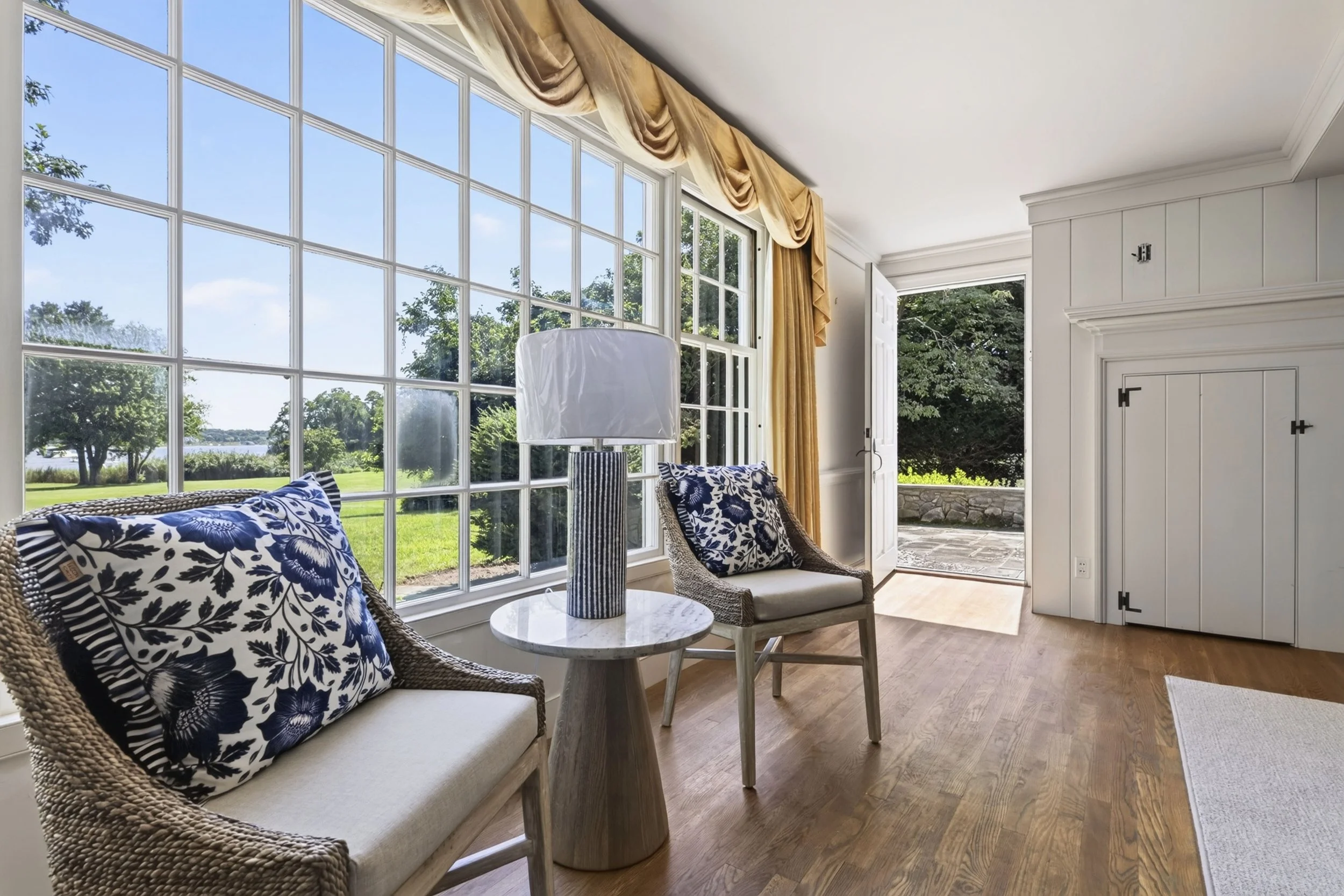 Living room with large window view of outdoor greenery and lake, wicker sofa with blue and white patterned pillows, round marble-top side table, and floor lamp, open door to outside with stone patio.