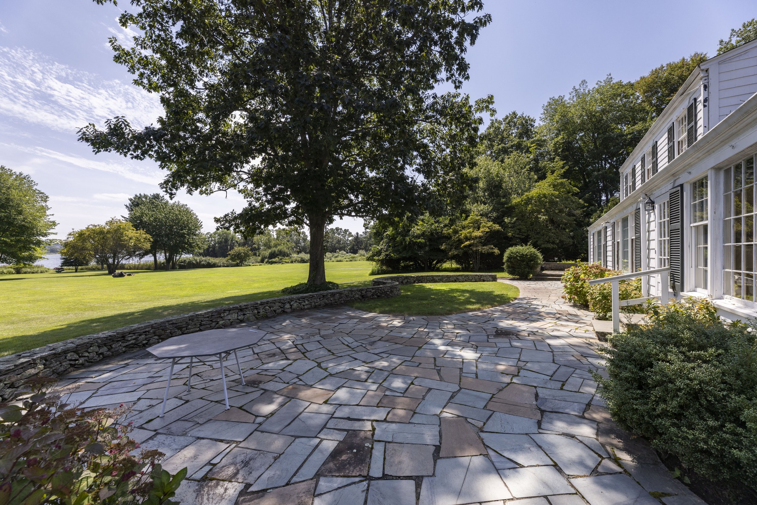 A house with large windows and black shutters, a stone patio, lush green lawn, large trees, and a lake in the background under a bright blue sky.