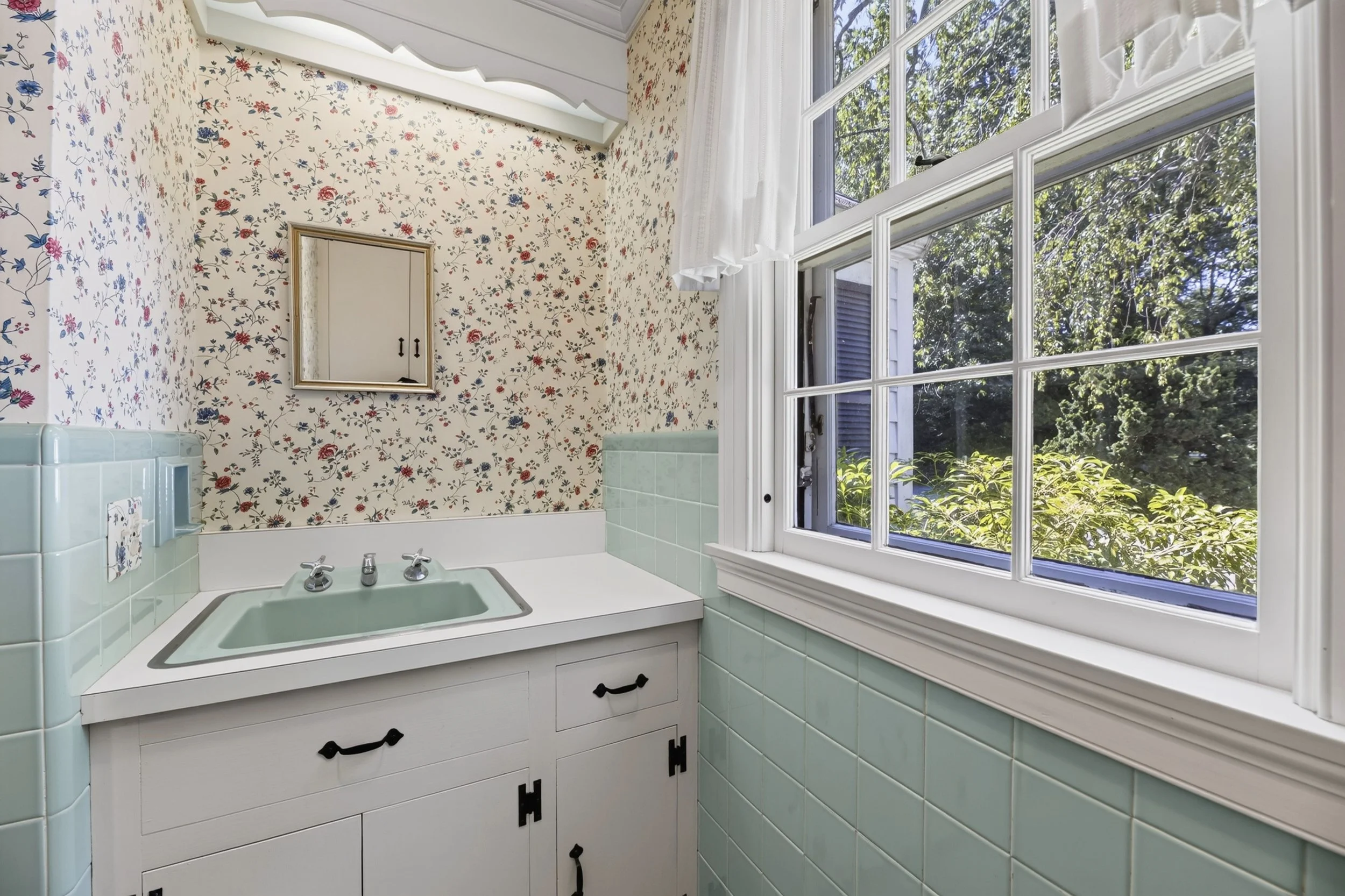 A vintage bathroom with a large window, floral wallpaper, a mint green bathtub, white cabinet with black handles, and a small mirror on the wall.