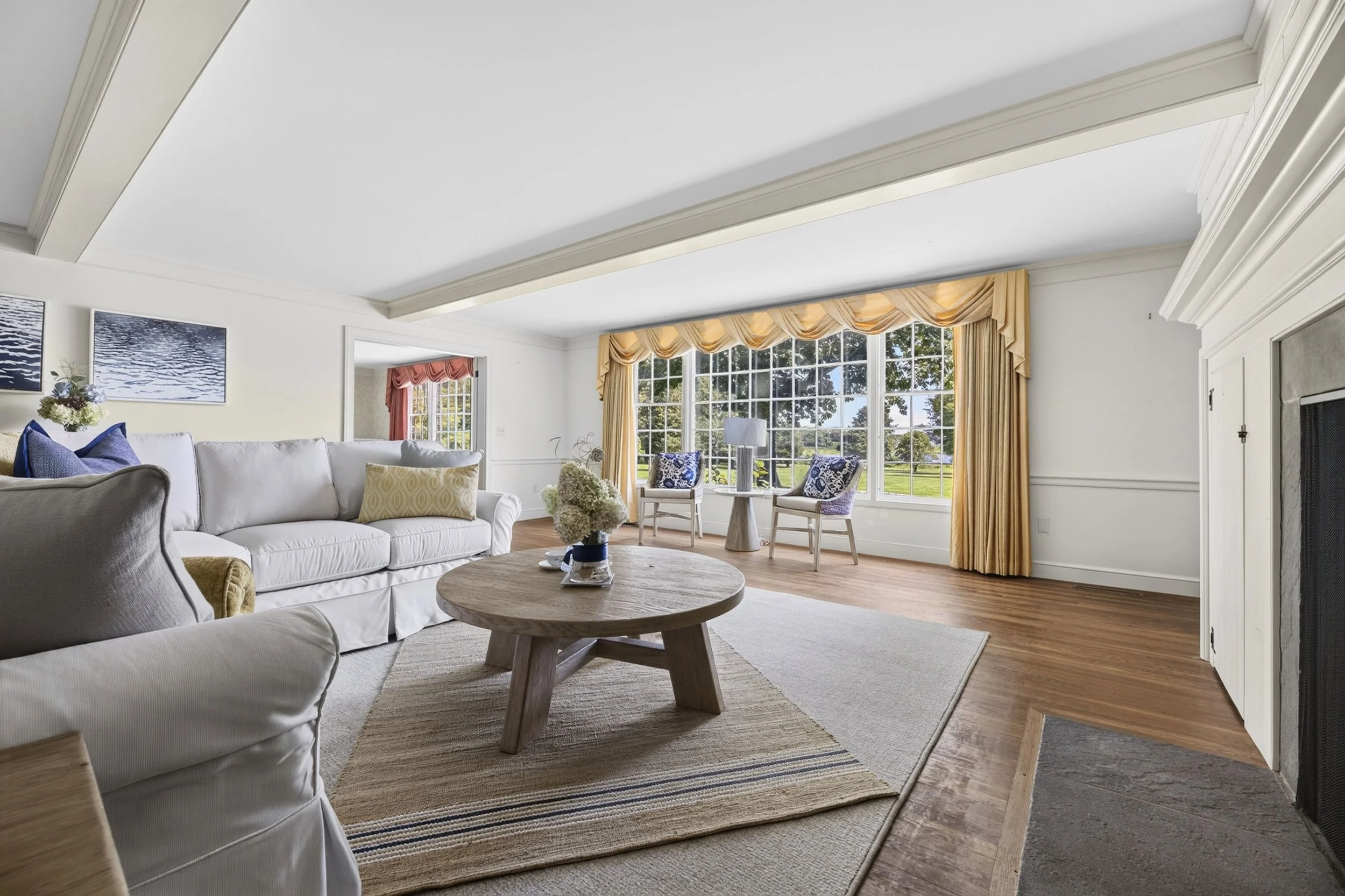 Living room with white sofa, armchair, wooden coffee table, large window with yellow curtains, and decorative pillows and artwork.