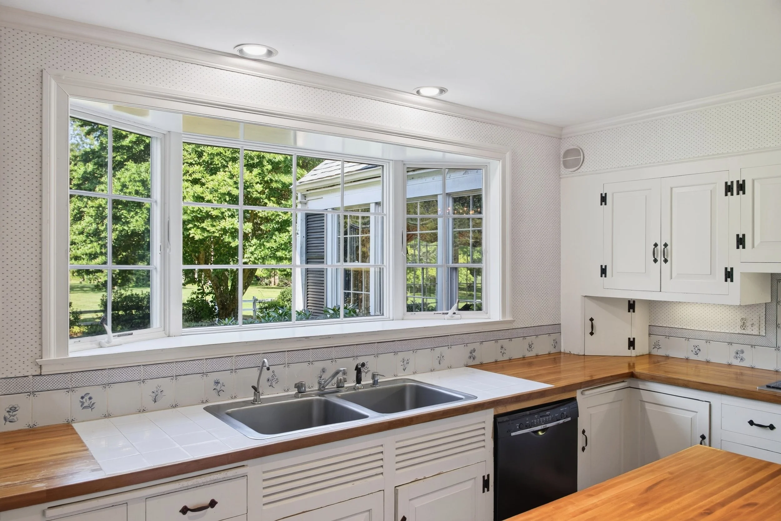 Kitchen with large window, white cabinets, marble tile countertop, stainless steel sink, wooden countertop accents, and view of green trees outside.