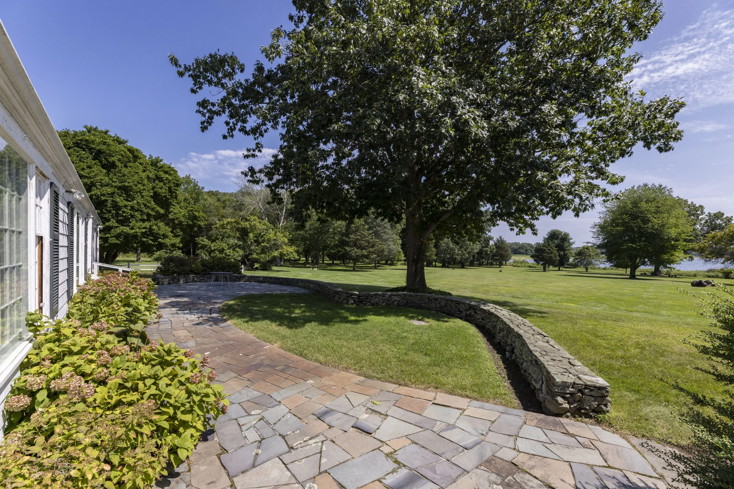 A landscaped backyard with a stone patio, flowering plants, a large tree, a grassy lawn, and a body of water in the background under a partly cloudy sky.